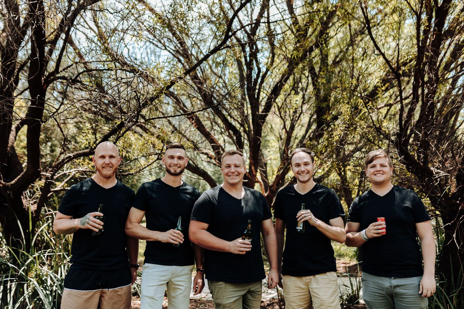 A group of five men in black t-shirts stand together outdoors under tree branches.