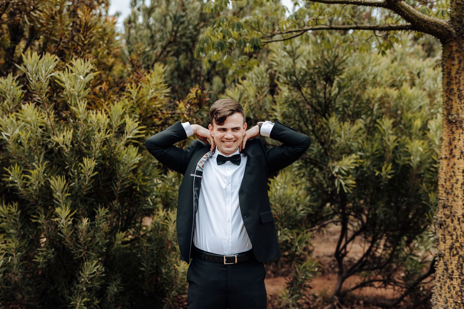 A person in a black tuxedo and bowtie adjusts their hair while standing among pine trees outdoors.