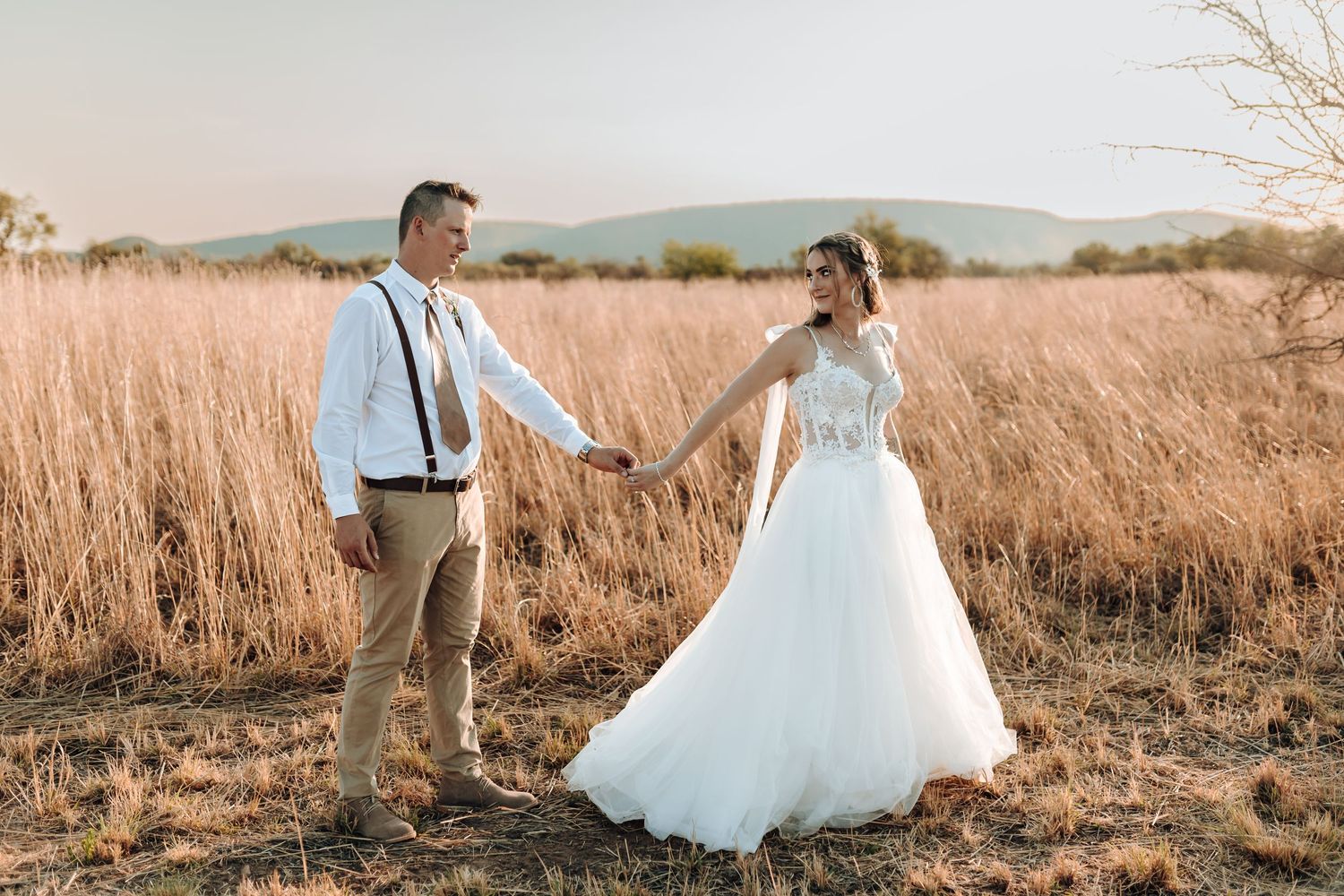 Wedding couple holds hands while walking through rustic field of tall grass with mountains in background.