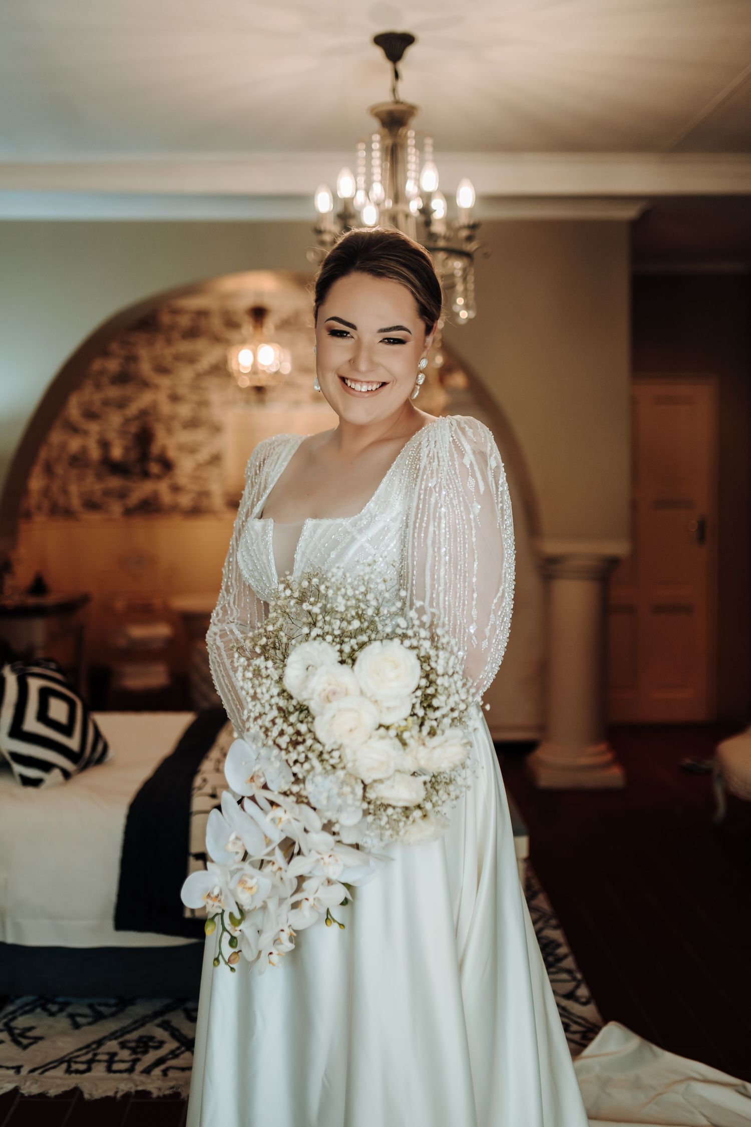 Elegant bridal portrait featuring a white wedding gown with sheer sleeves, crystal chandelier, and white floral bouquet.