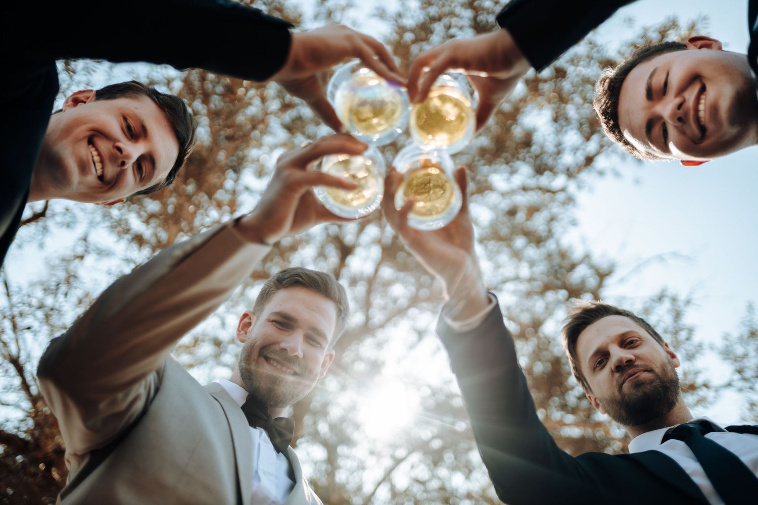 Wedding party toasts with champagne glasses from below against sunlit sky.