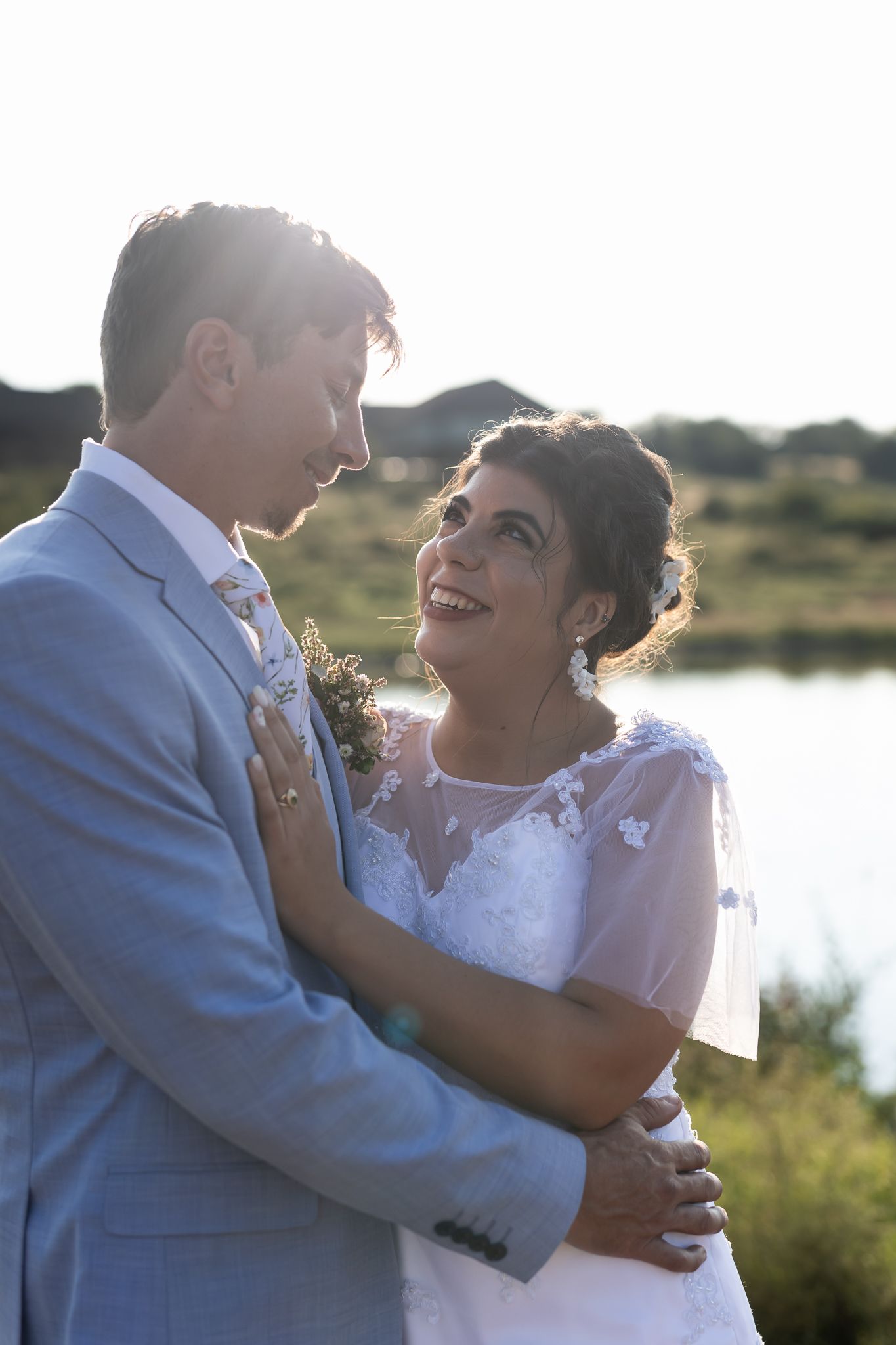 A romantic wedding couple shares a tender moment by a lake during golden hour sunset.
