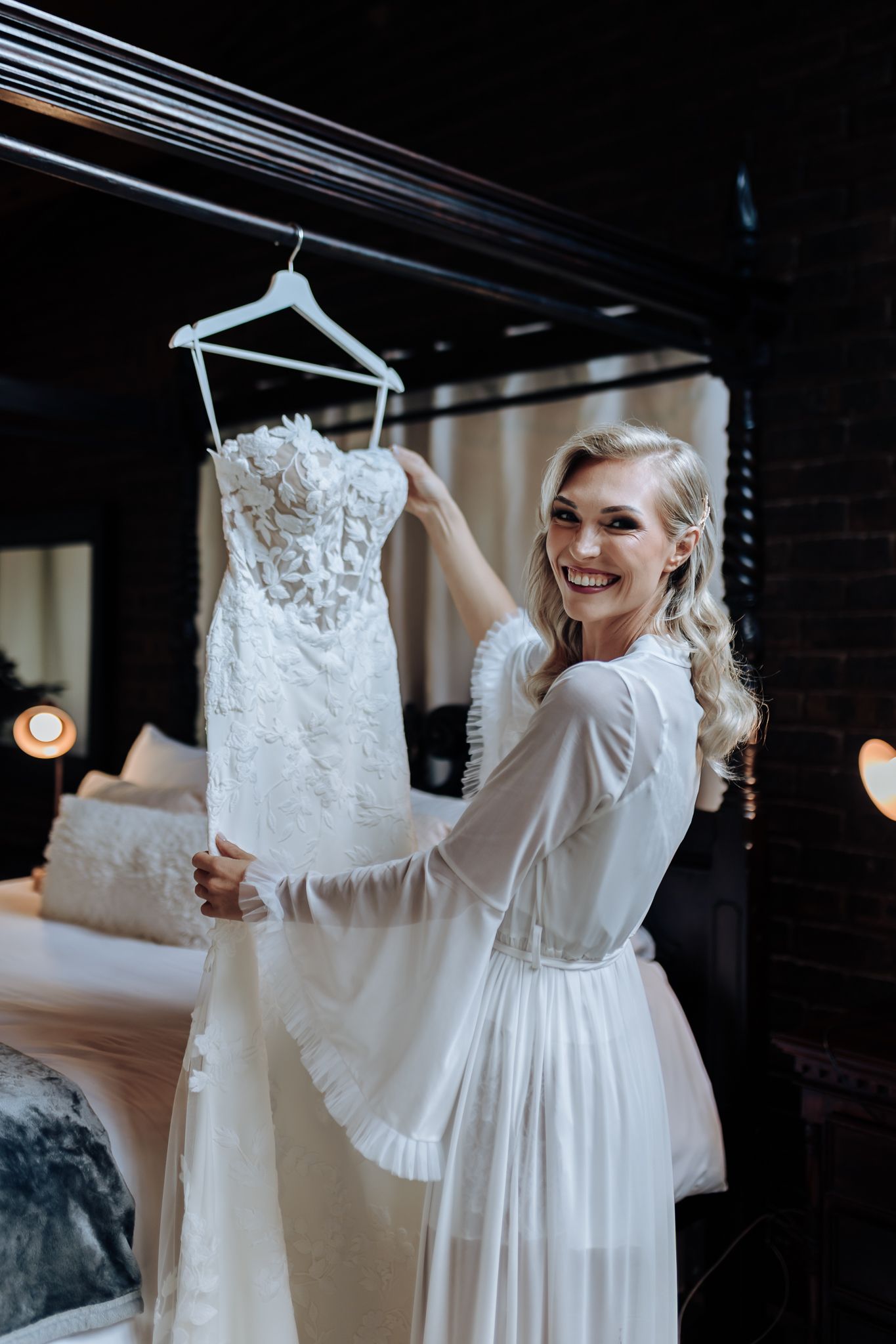 Bride in white robe admiring lace wedding dress hanging in rustic getting-ready room.