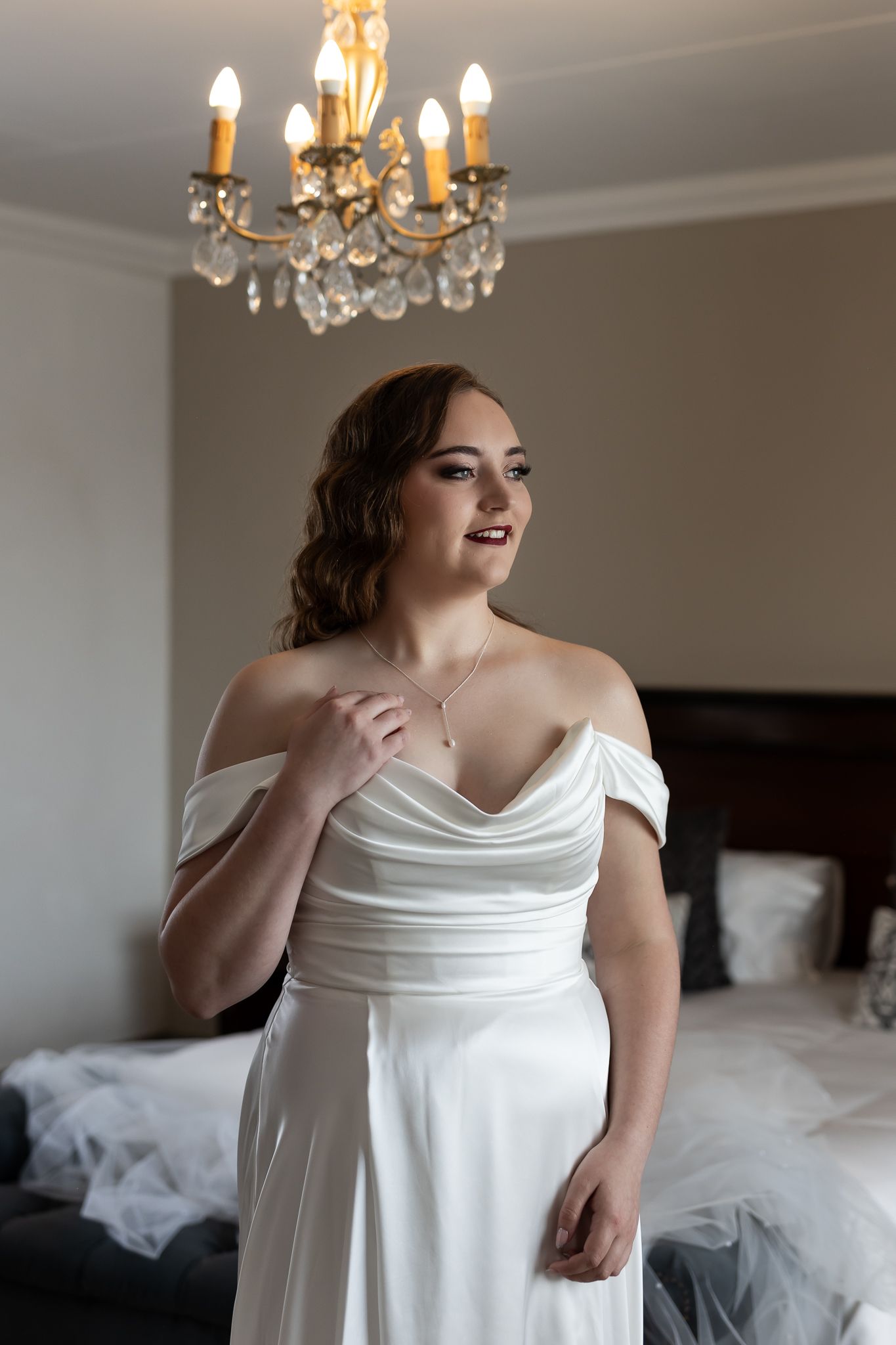 Elegant bridal portrait under crystal chandelier in white off-shoulder gown.