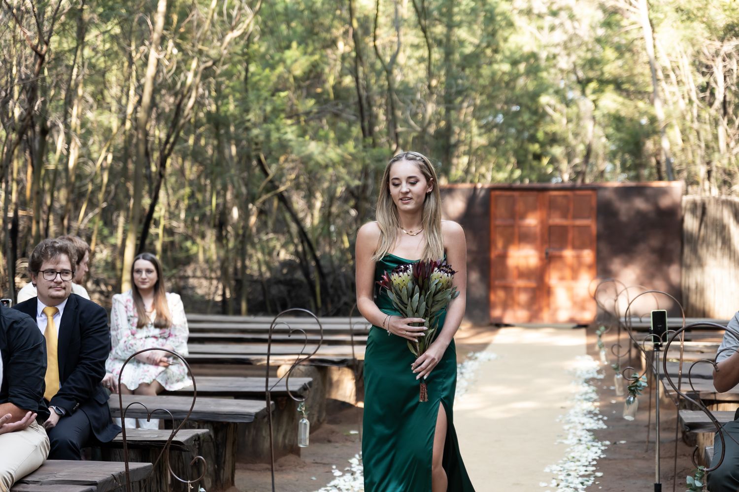 Bridesmaid in emerald dress walking down wooden aisle in forest setting.