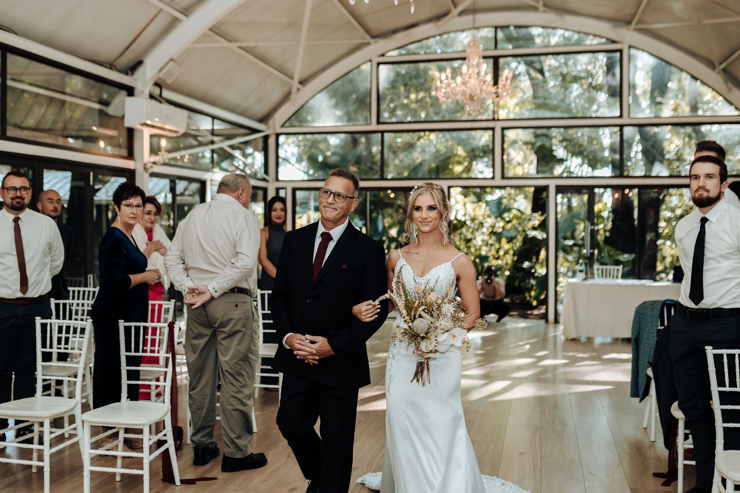 Wedding ceremony procession in a bright venue with arched windows and white chairs.