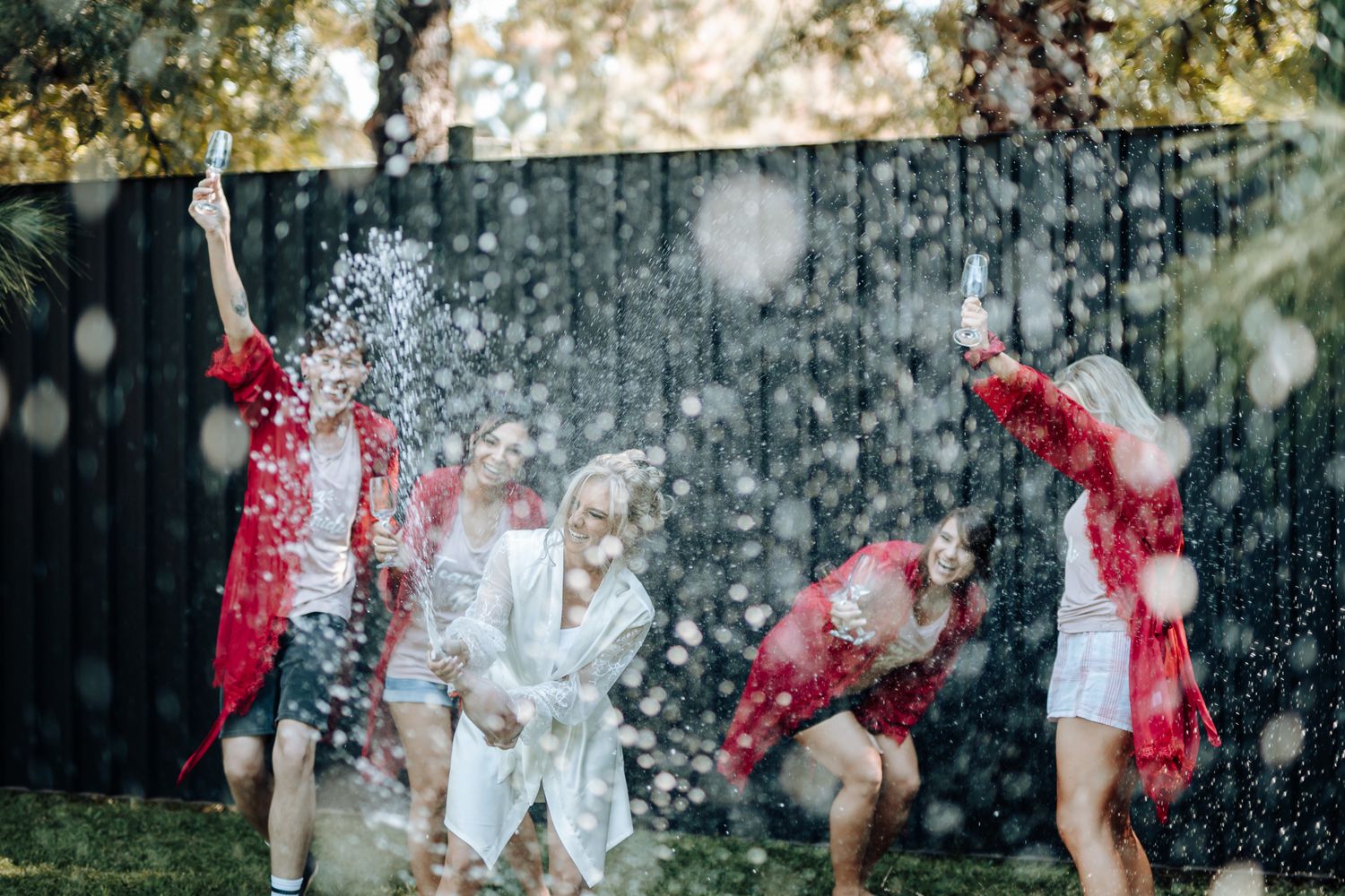 Friends in red and white outfits celebrating with champagne spray in a backyard.