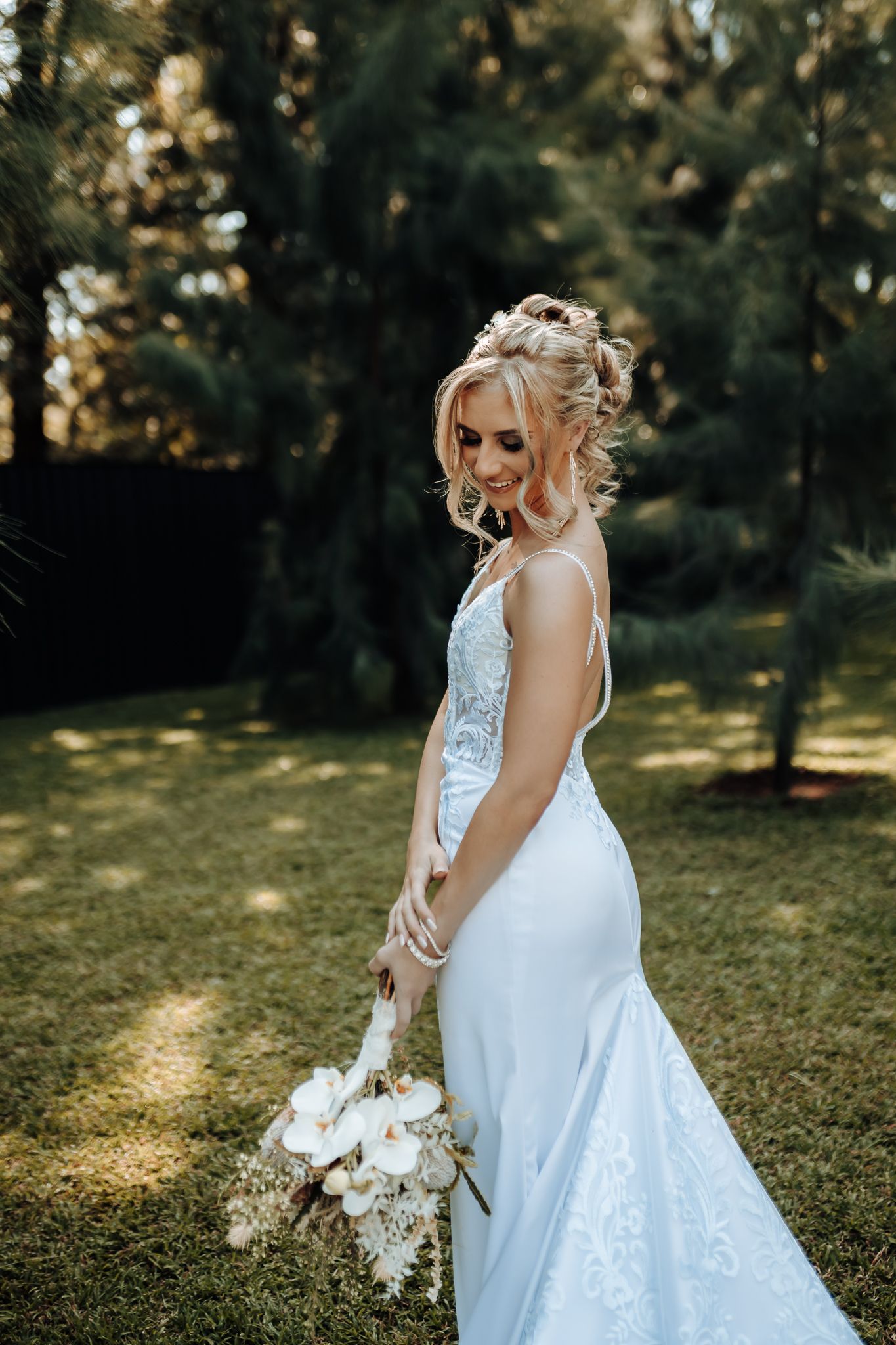 Elegant bride in a fitted lace wedding dress poses outdoors holding a white bouquet in a garden setting.