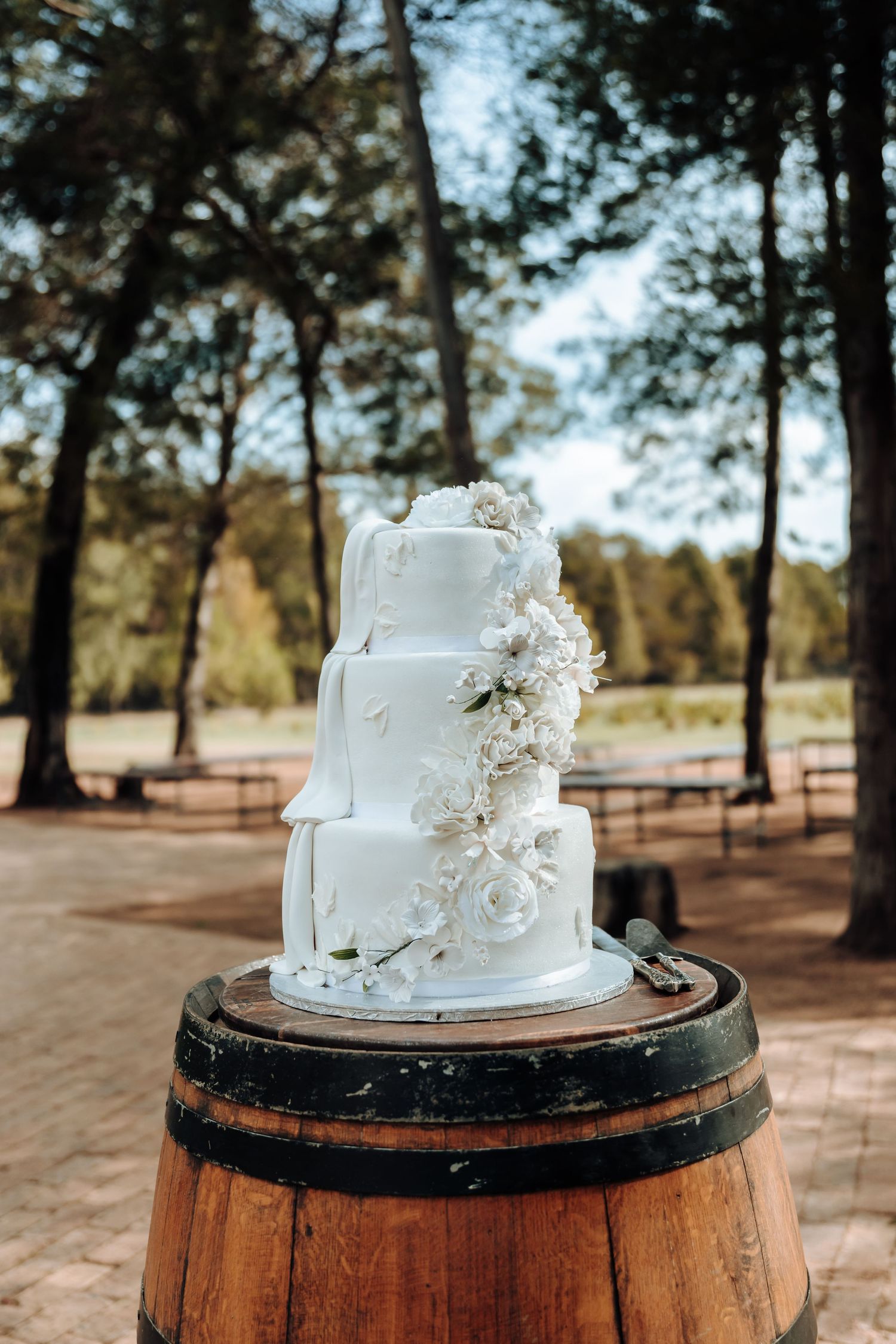 Elegant white wedding cake with delicate floral details displayed on rustic wooden barrel outdoors.