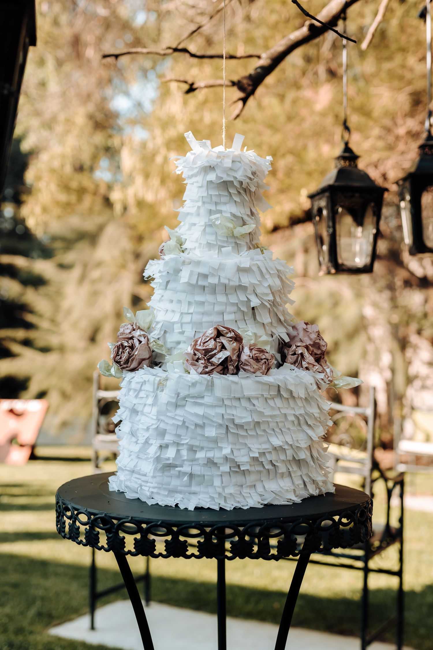 An elegant white ruffled wedding cake with brown floral accents displayed on a black stand outdoors.