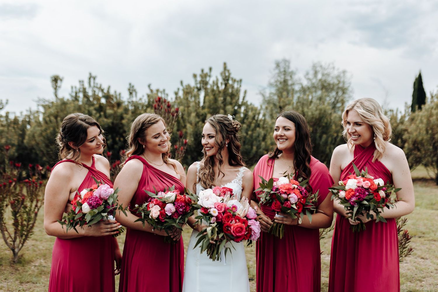 Wedding party poses outdoors with bridesmaids in red dresses holding colorful floral bouquets against natural backdrop.