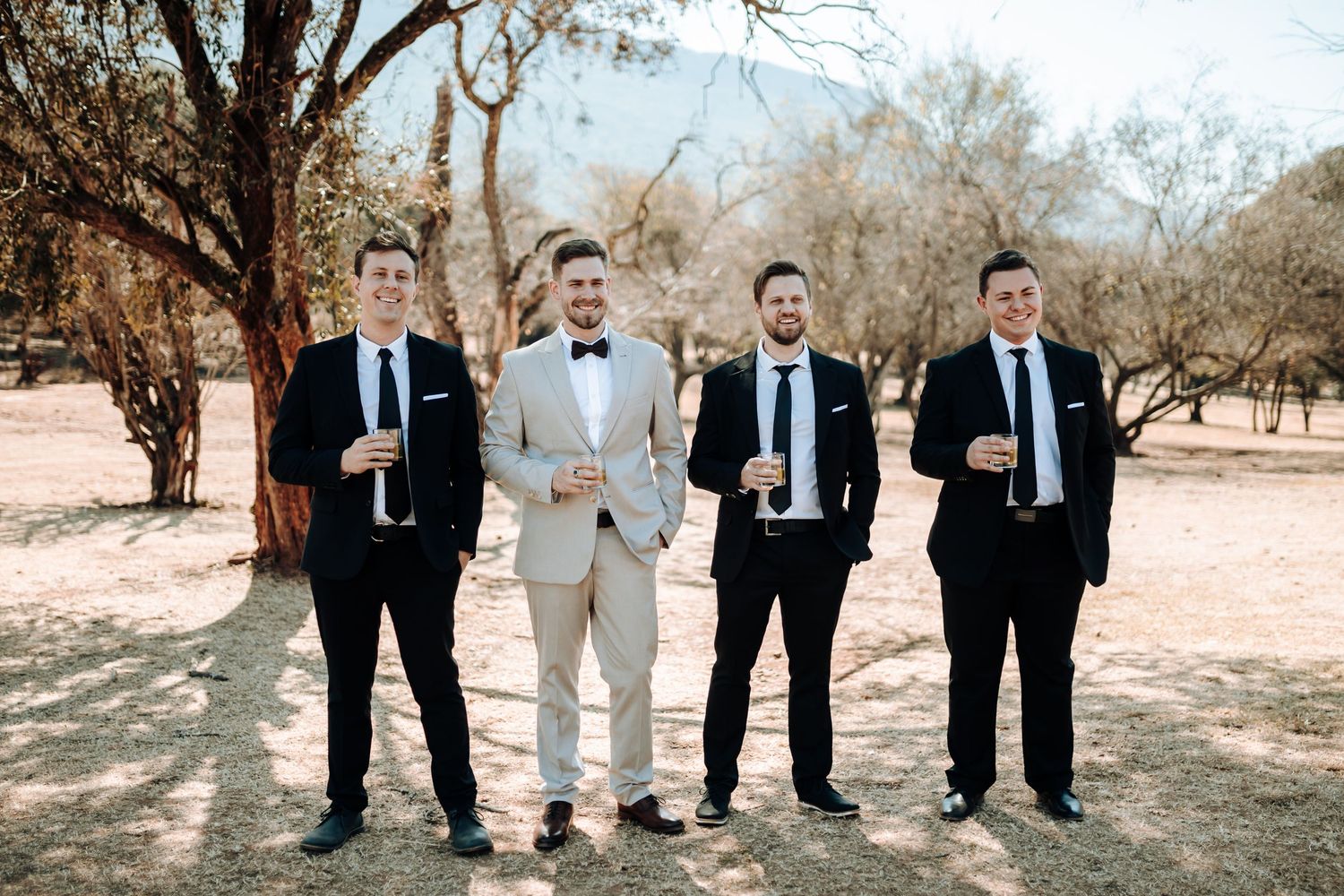 Four men in formal attire pose together in a rustic outdoor setting with desert landscape.