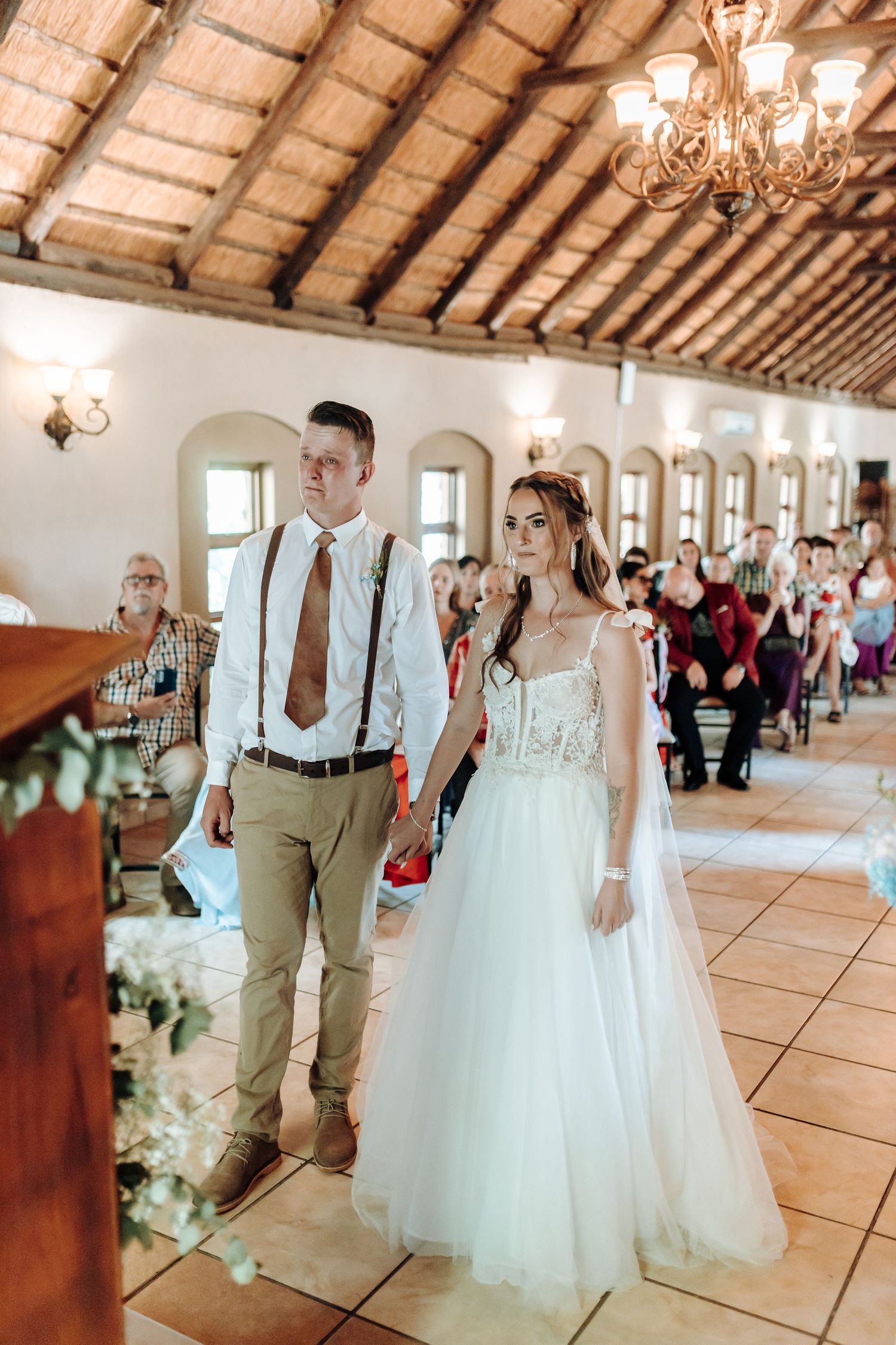Couple stands at altar in rustic venue with wooden beam ceiling and elegant chandeliers.