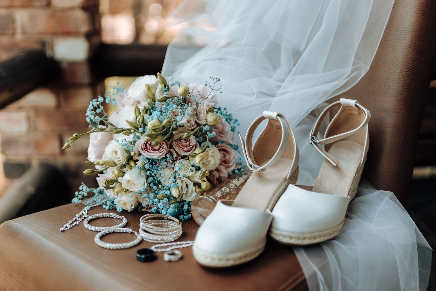 Wedding details including bridal bouquet with succulents, white heels, and jewelry on brick surface.