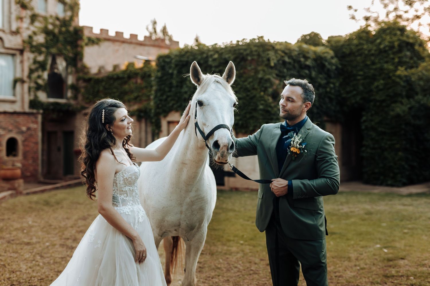 Couple in wedding attire interacting with a majestic white horse by an ivy-covered castle wall.