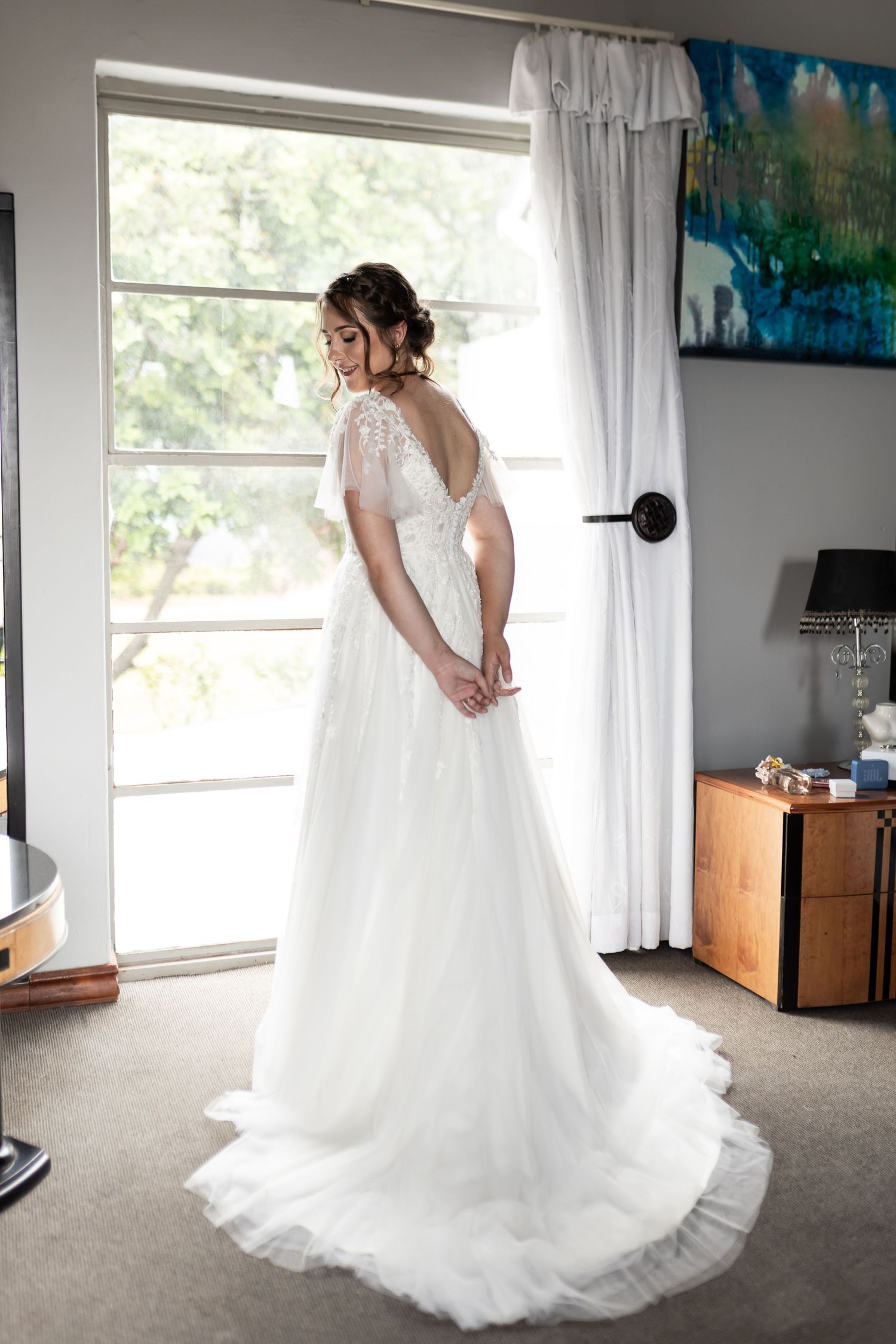 A stunning white wedding gown with lace details and a flowing train shown from behind near a bright window.