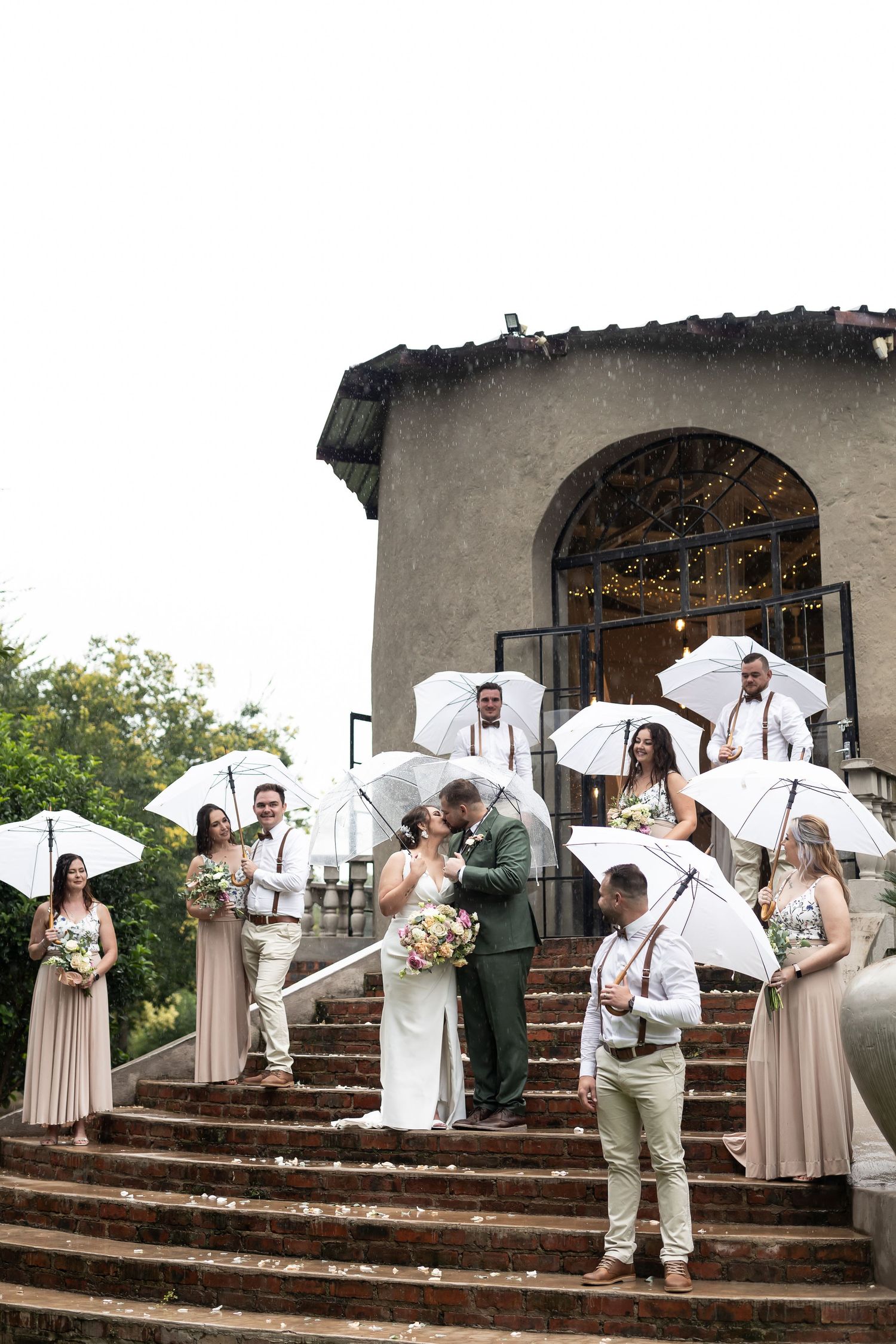 Wedding party poses with white umbrellas on stone steps of rustic venue.