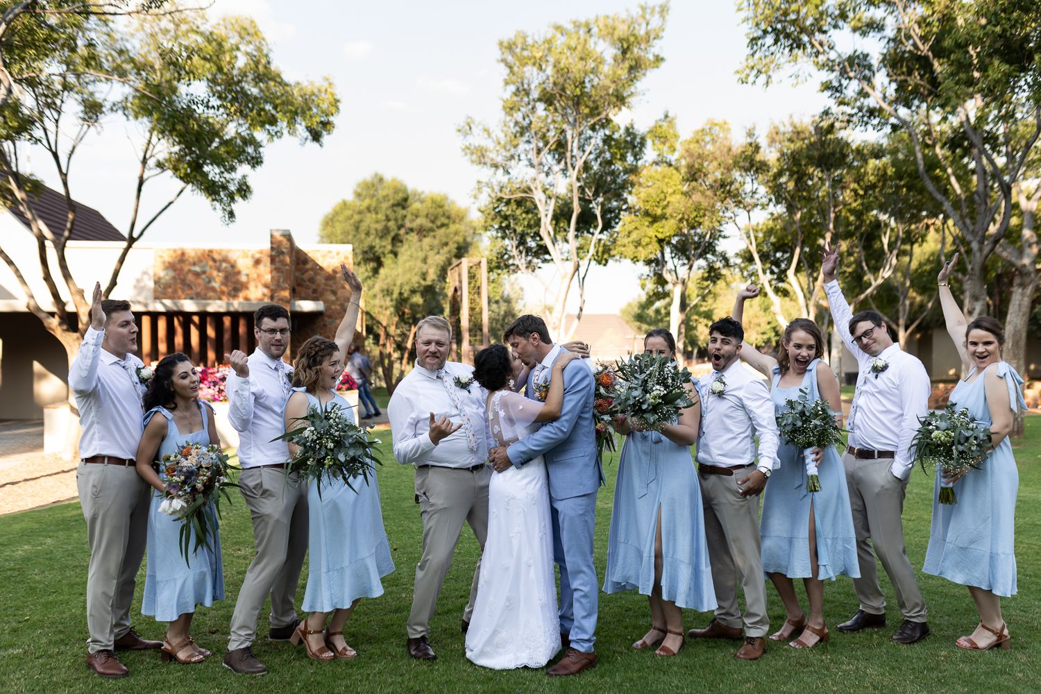 Wedding party cheering and celebrating with newlyweds on lawn in coordinated attire.