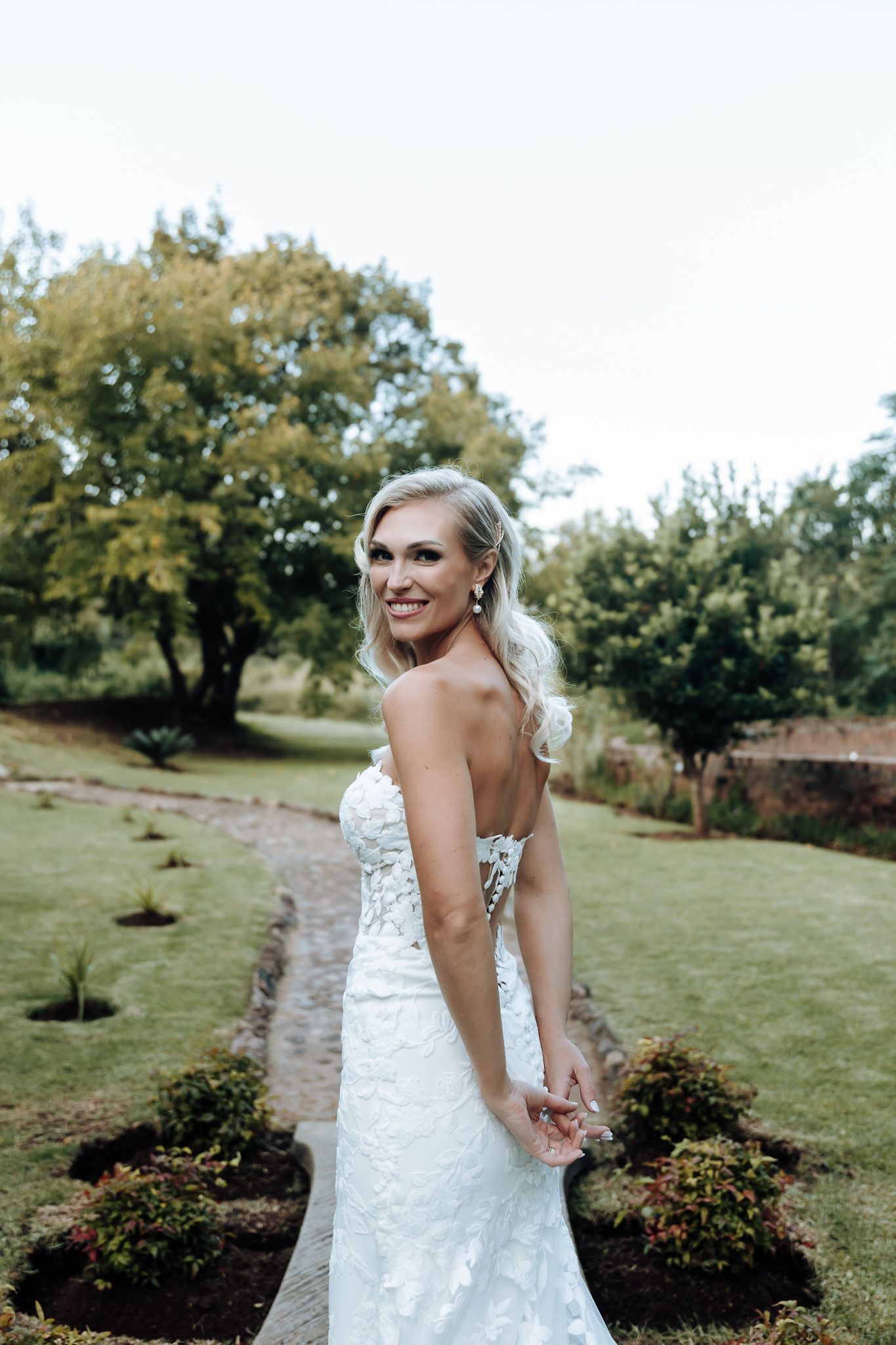 A stunning lace wedding dress is showcased in an outdoor garden setting with blooming flowers and trees.