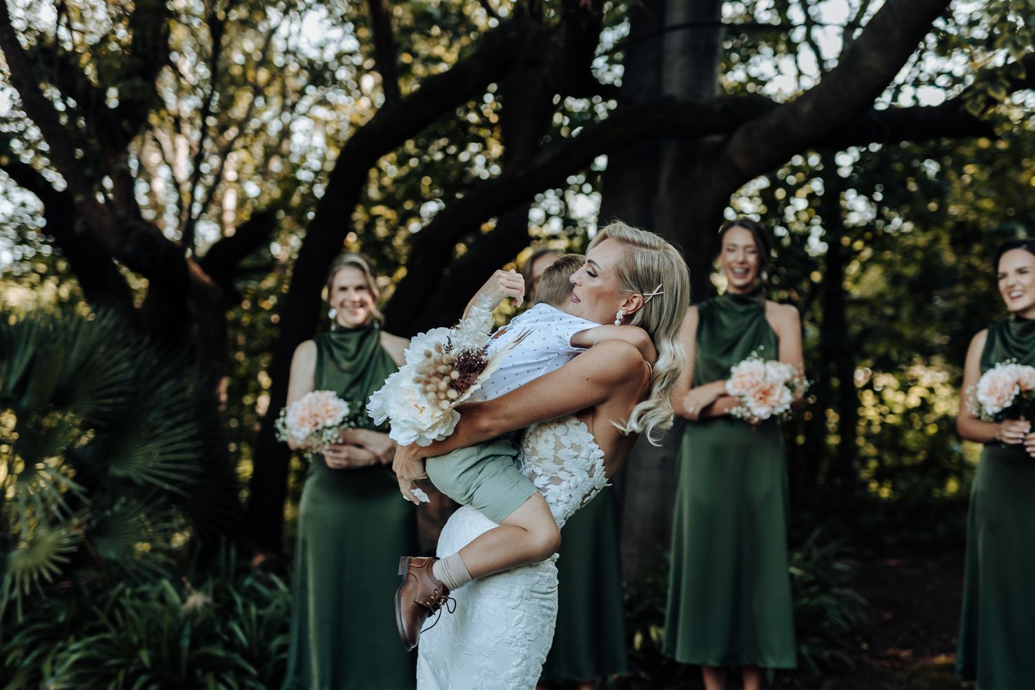 Bride in lace dress embraces child while bridesmaids in emerald dresses stand in outdoor garden setting.