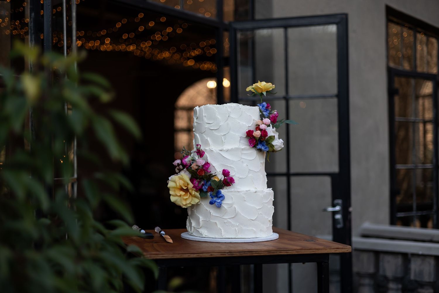 A white three-tiered wedding cake decorated with fresh colorful flowers sits on a wooden table outdoors at dusk.