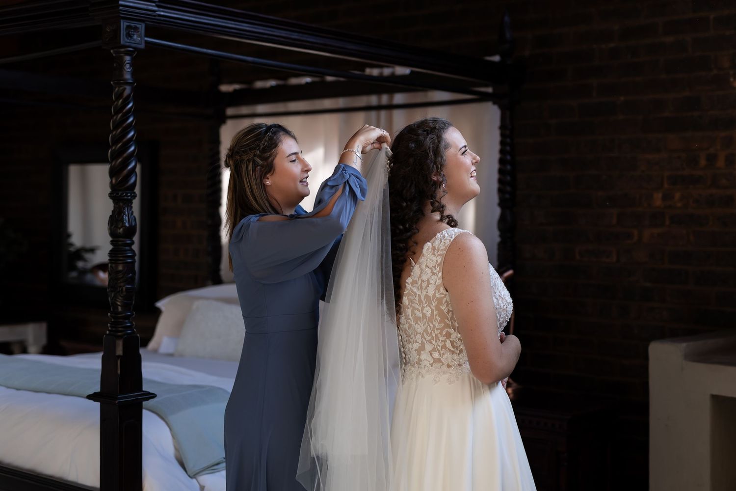 An elegant bride has her veil adjusted during a wedding preparation moment against a dark brick wall.