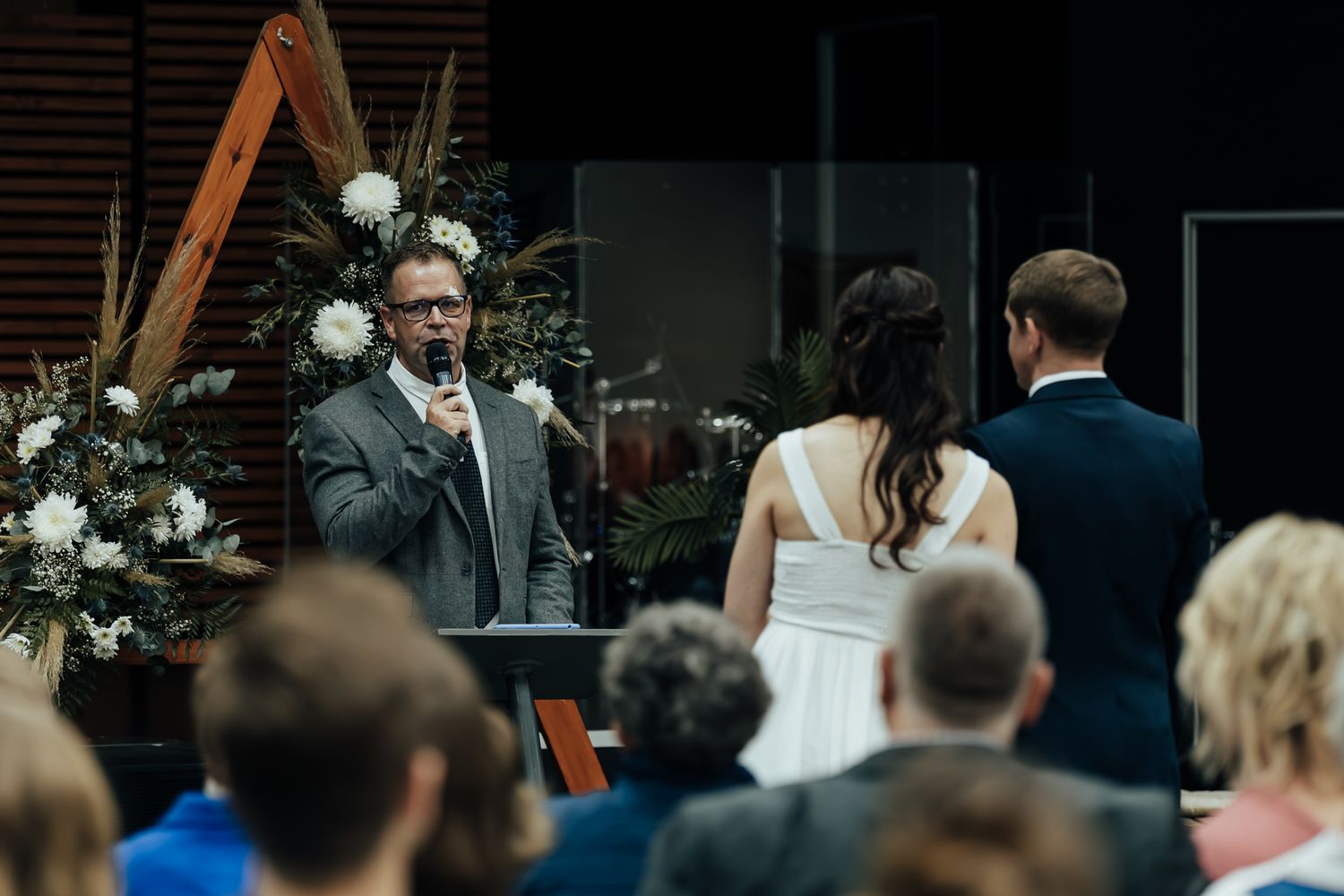 A wedding ceremony in progress with an officiant speaking at a wooden arch decorated with white floral arrangements.