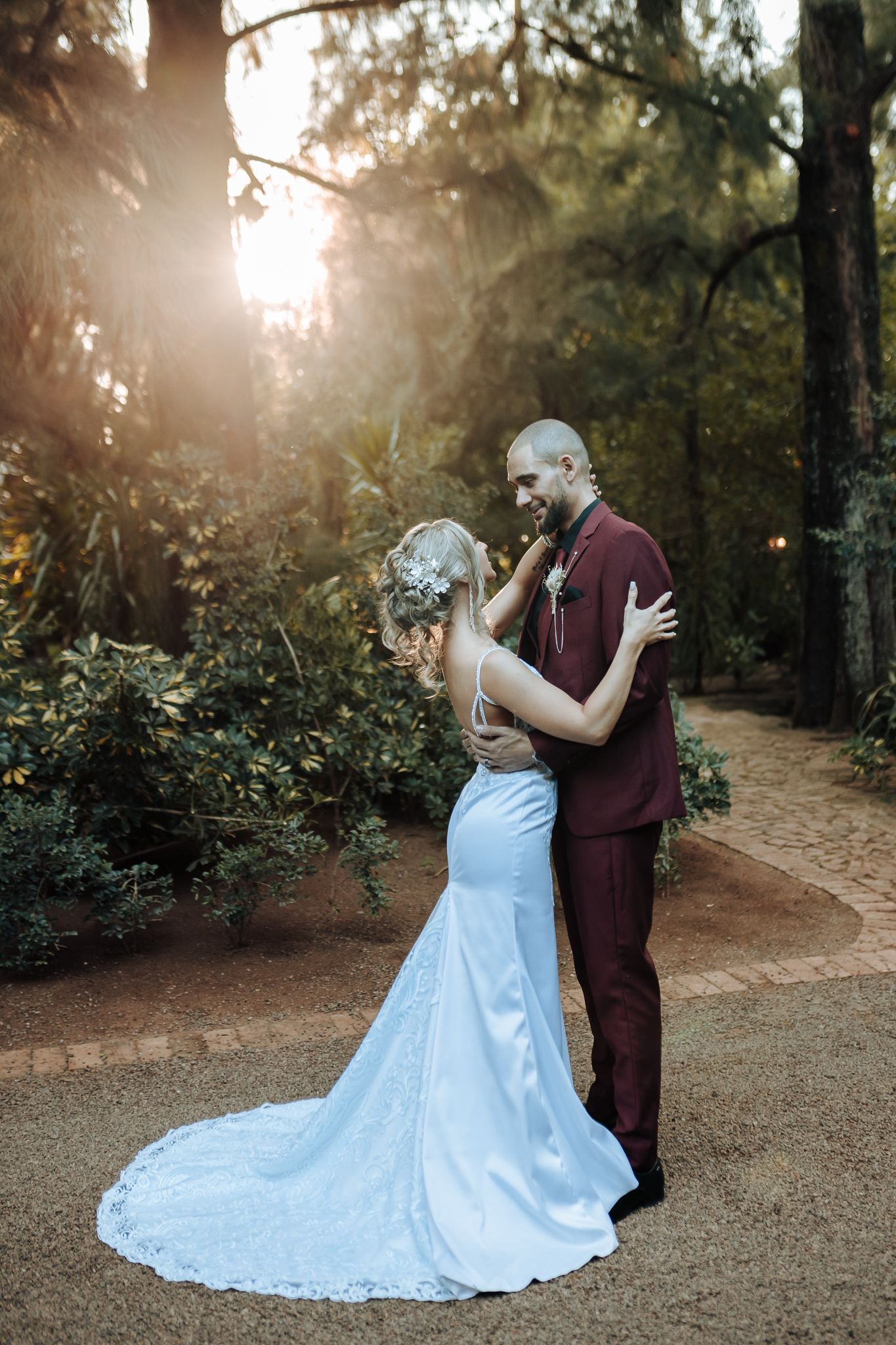 Couple shares intimate dance moment in elegant wedding attire with sunlight through trees.