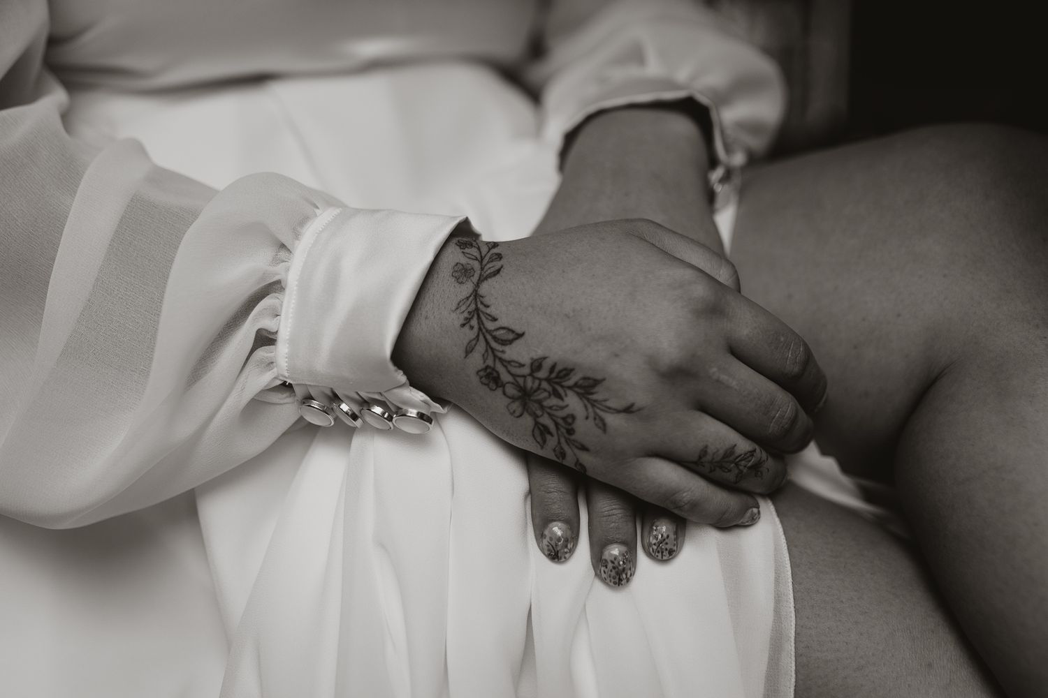 Close-up black and white photo of hands with floral tattoo and bracelet against white fabric.