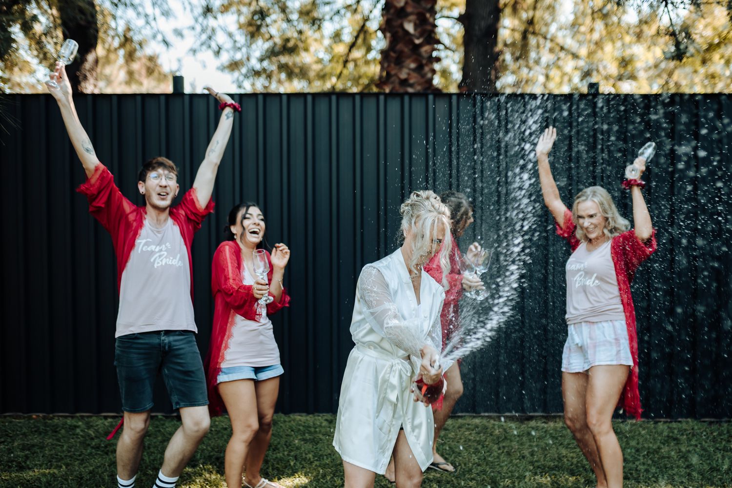 Group celebrating with champagne spray outdoors wearing red and white outfits.