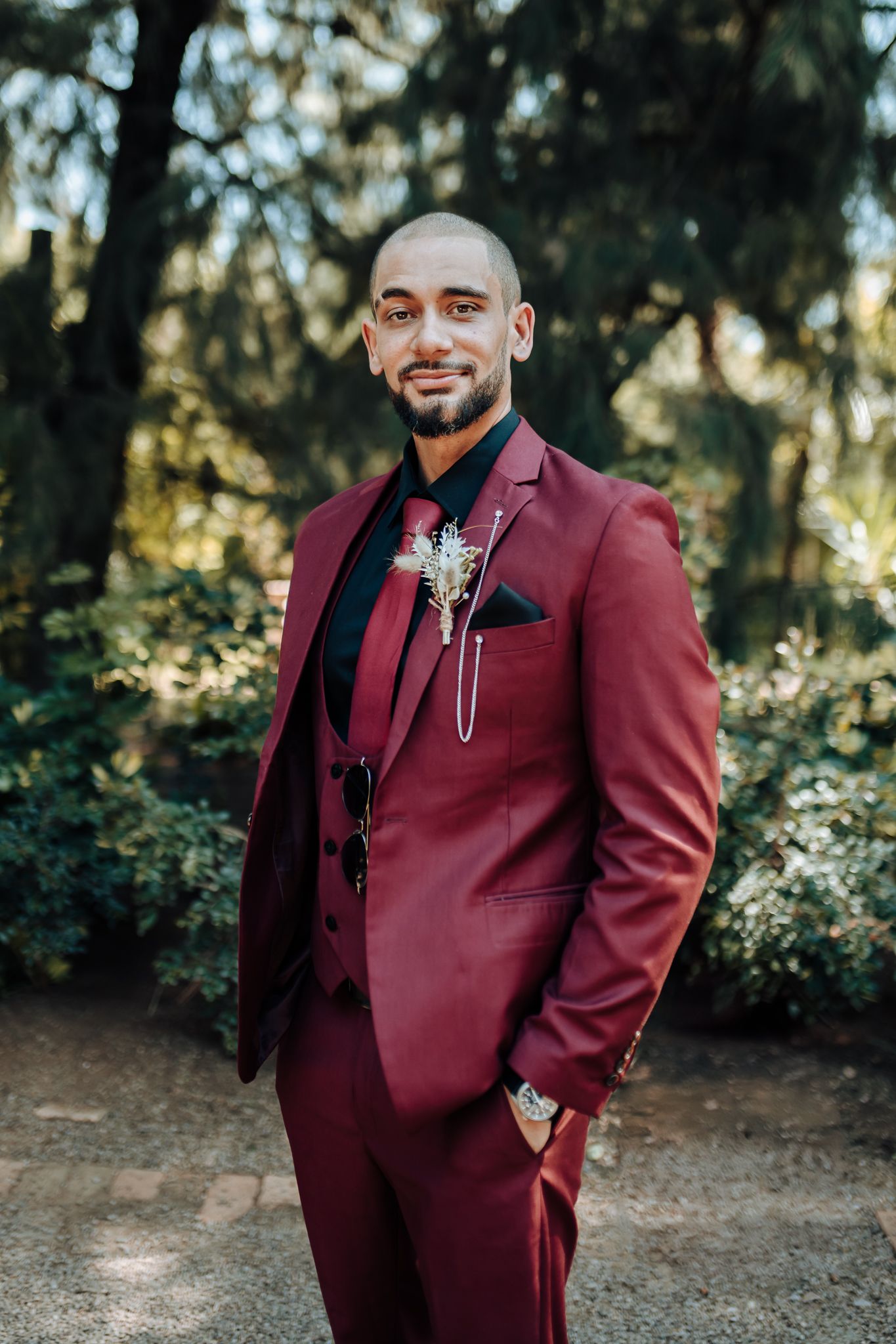 Groom in burgundy suit with boutonniere poses in outdoor garden setting.