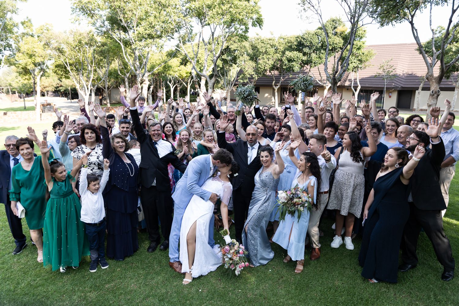 Large wedding party group photo with guests cheering and celebrating on sunny lawn.