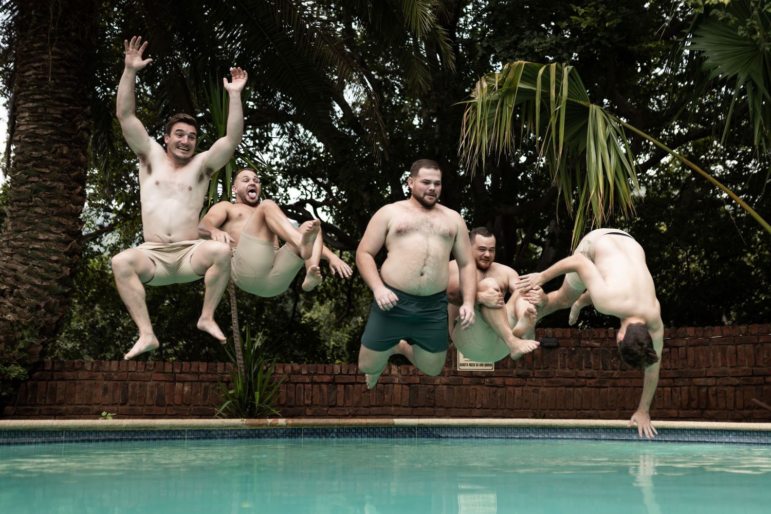 Groomsmen jumping into a swimming pool in synchronized formation against a brick wall backdrop.