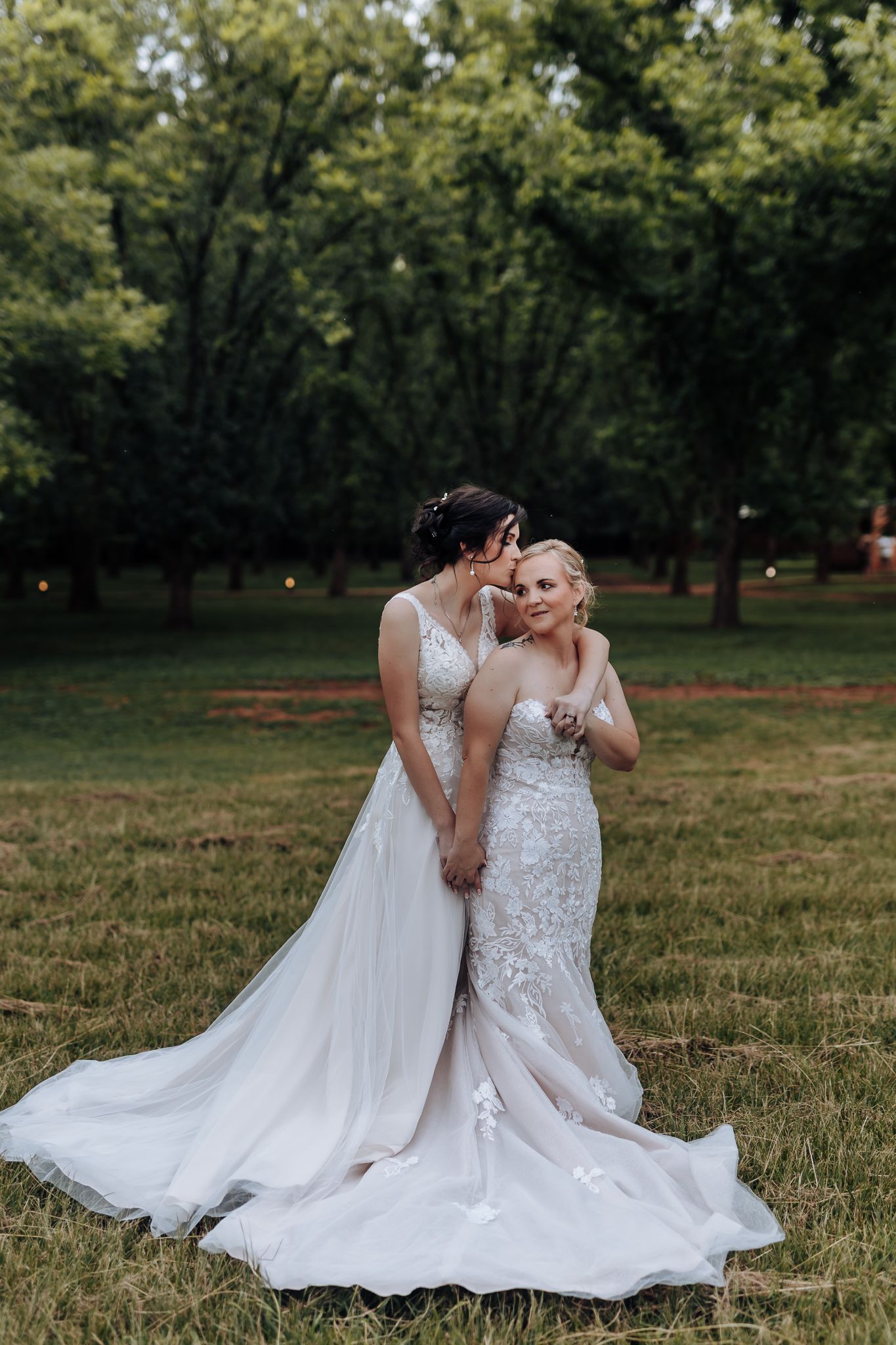 Two brides in flowing white gowns pose together on grass with trees in the background.