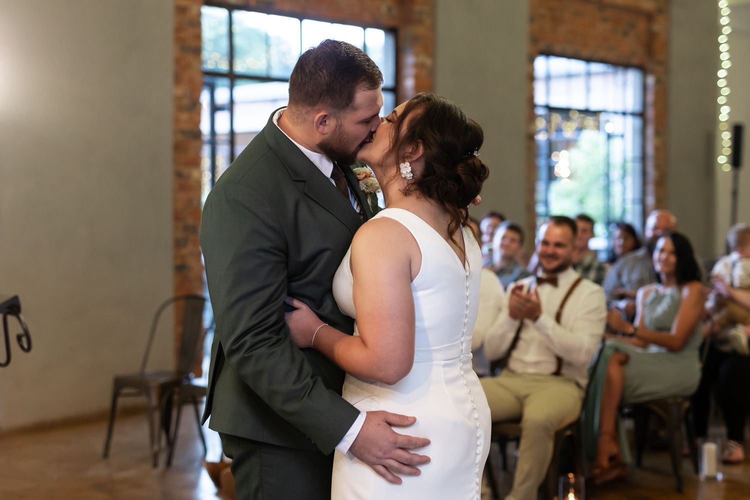Couple shares intimate first dance moment in rustic brick-walled venue with wedding guests watching in background.