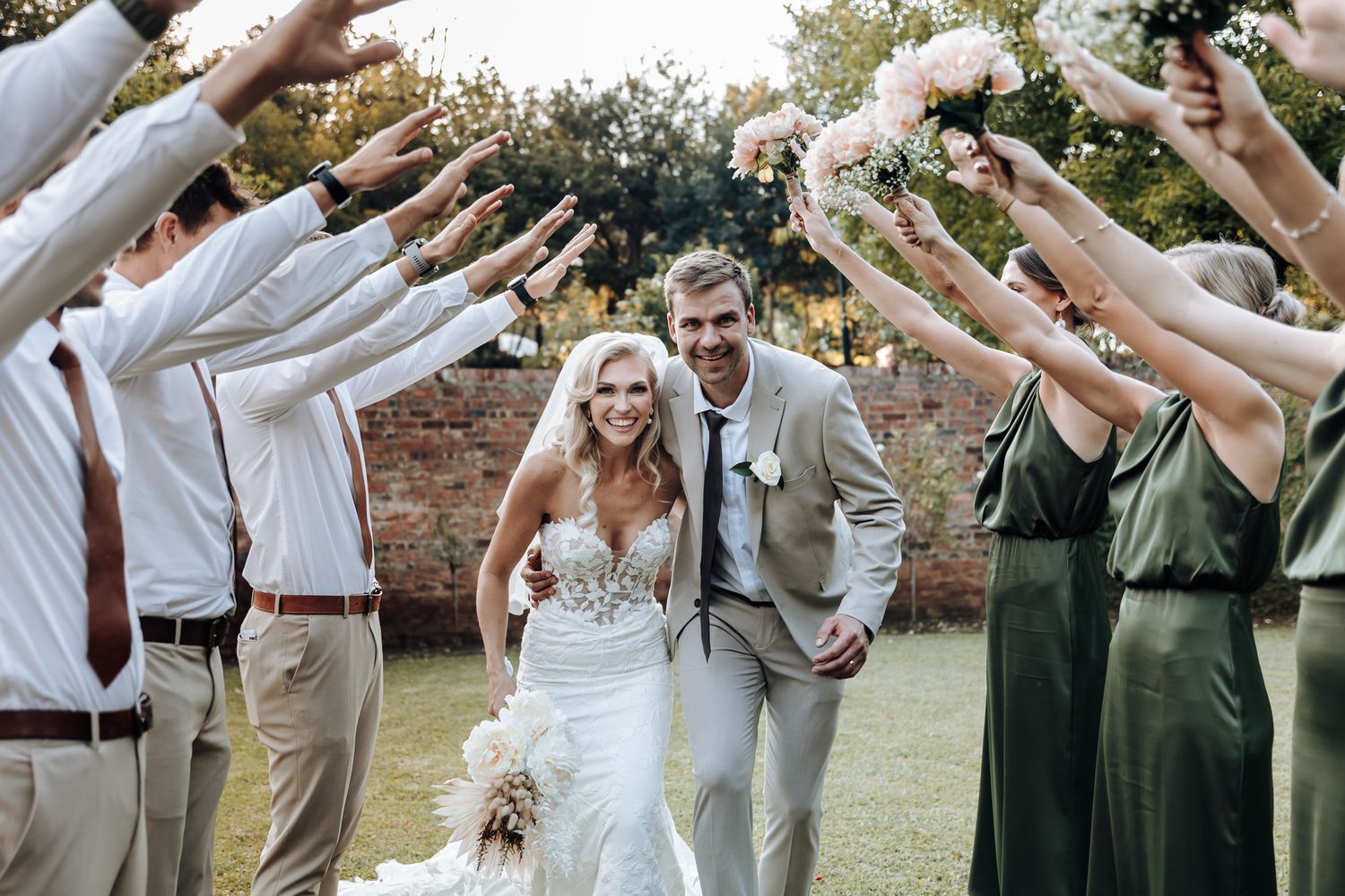 Wedding guests form tunnel with raised arms as newlyweds walk through during outdoor celebration.