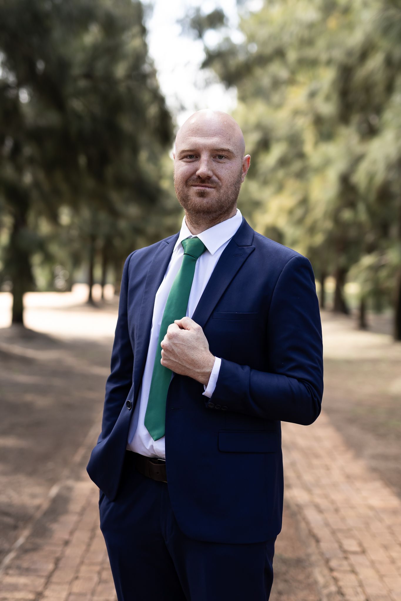 Formal portrait of man in navy suit with emerald tie standing on brick pathway.