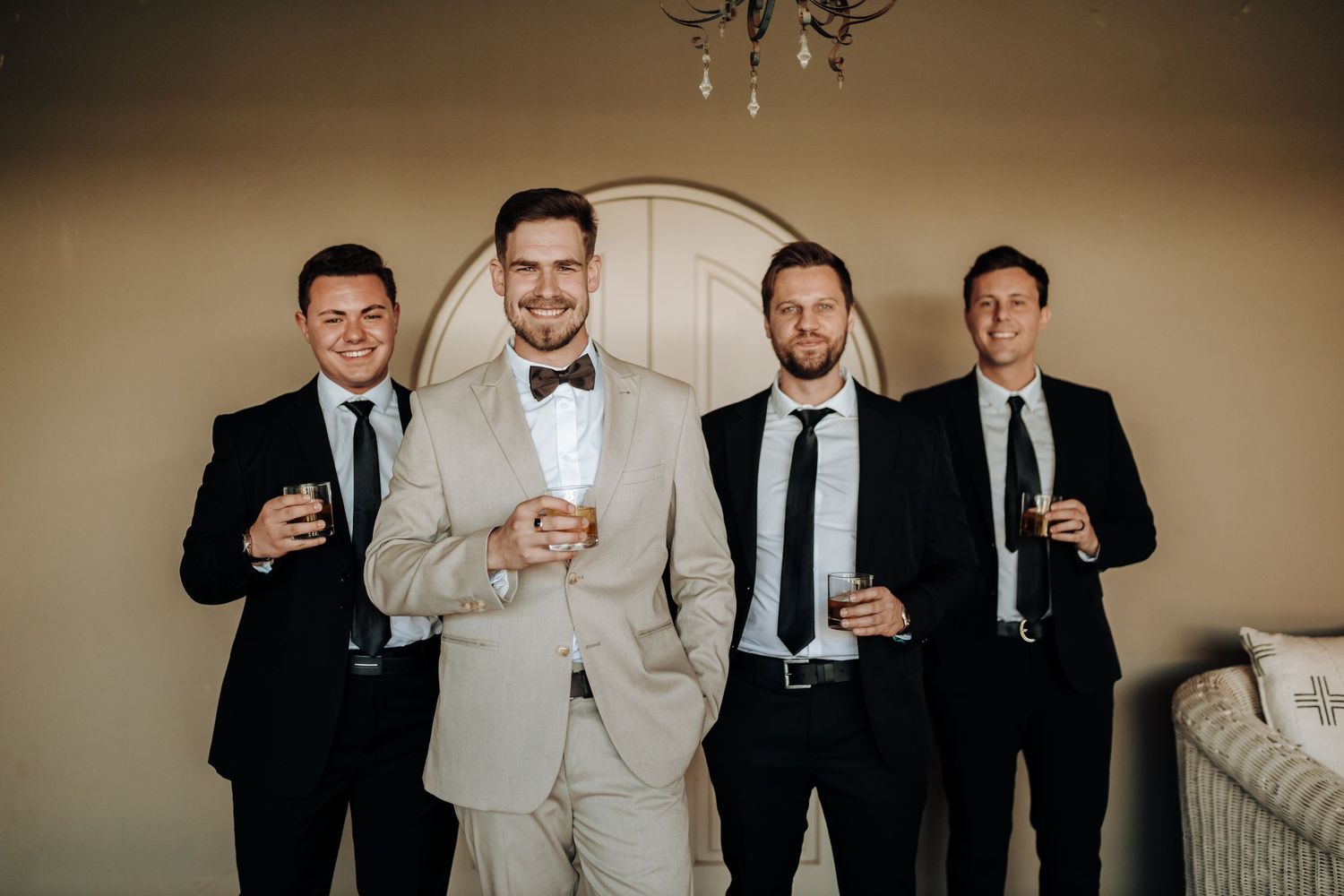 Wedding party of groomsmen in black suits surrounding groom in light beige suit holding drinks.