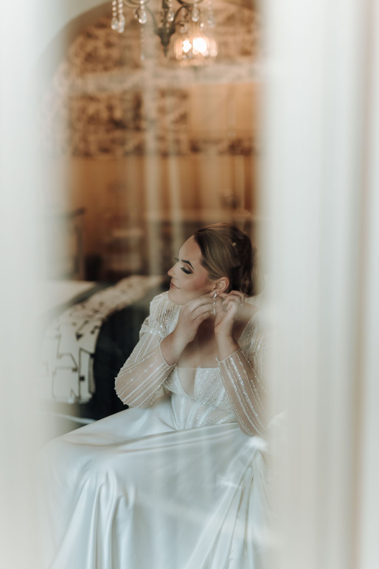 Dreamy portrait of bride putting on jewelry, viewed through sheer curtains.