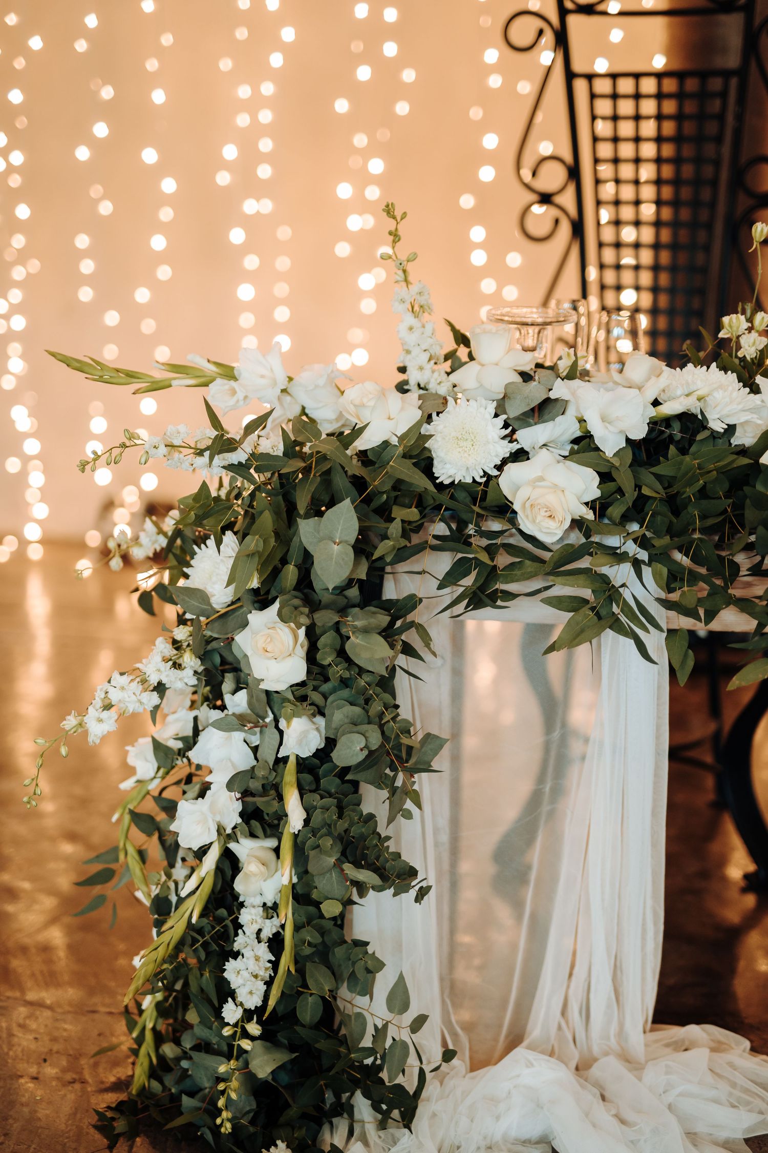 White floral arrangement with cascading greenery against twinkle light backdrop.