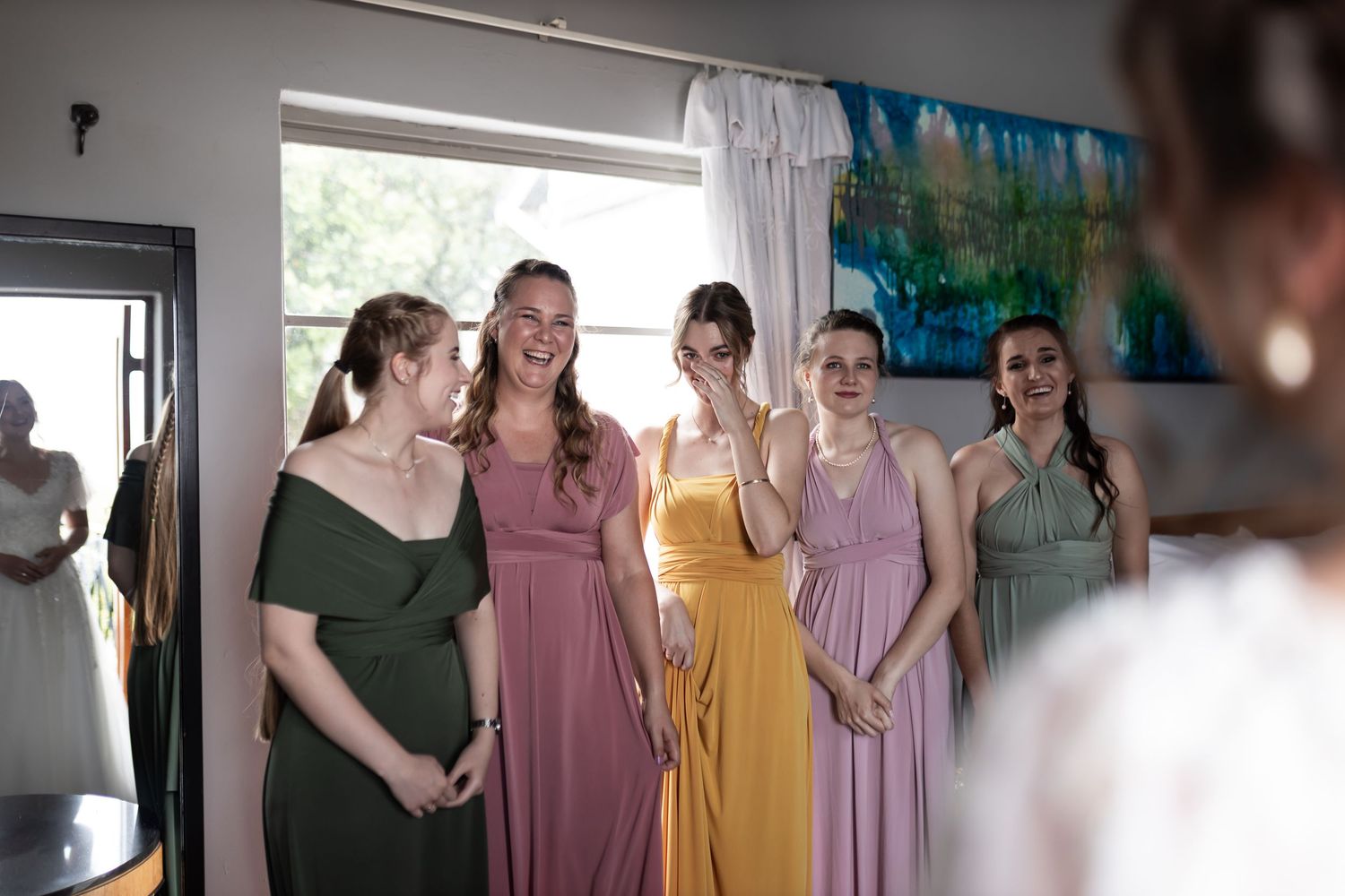 Bridesmaids in jewel-toned dresses share a joyful moment together in front of a window.