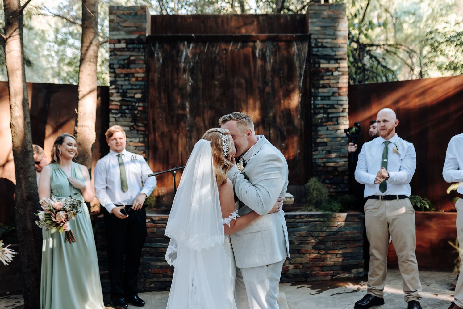 A wedding ceremony takes place outdoors with a rustic stone fireplace backdrop and members of the wedding party in sage green.