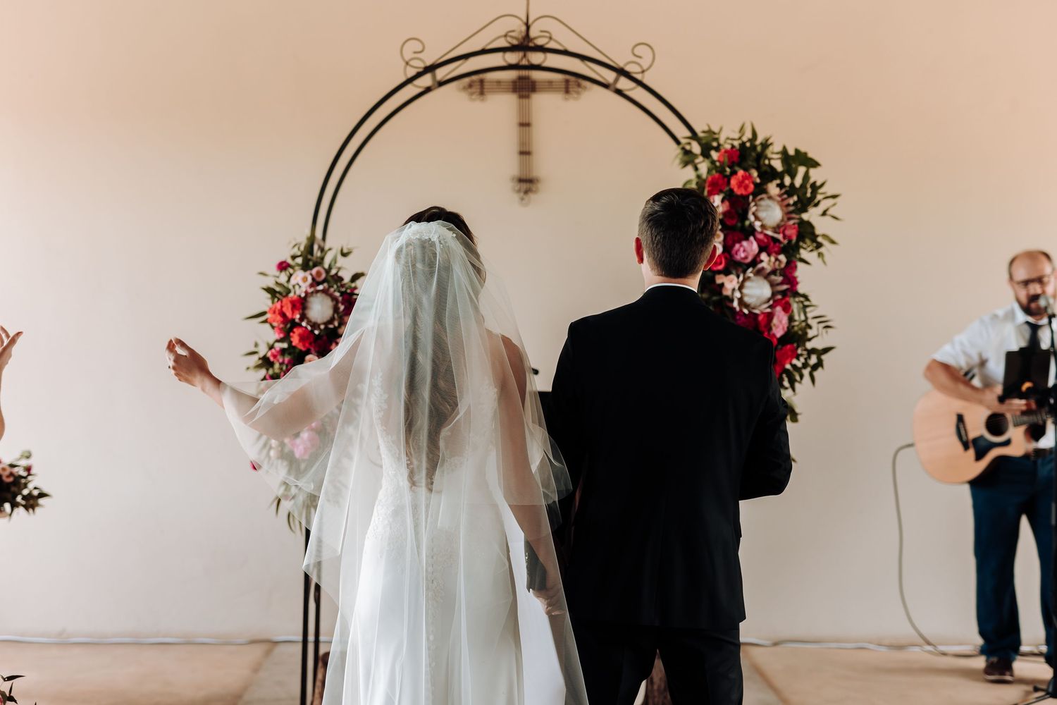 Ceremony under decorative metal arch adorned with pink and red florals.