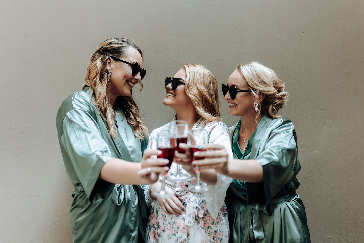 Friends in mint green robes and sunglasses toast with wine glasses outdoors.