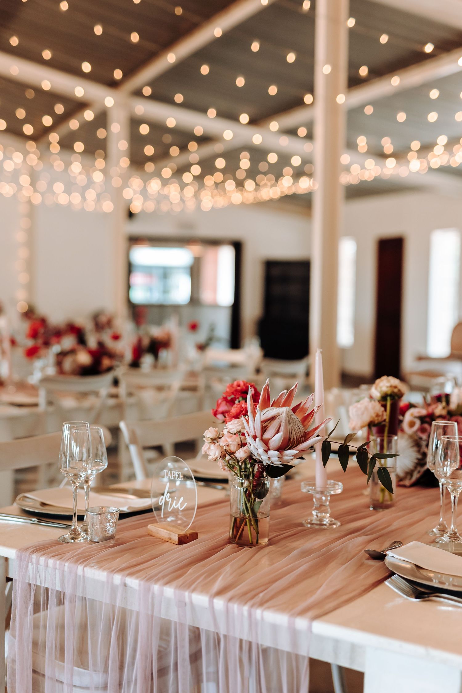 Wedding reception table with string lights overhead and pink floral centerpieces on wooden tables.