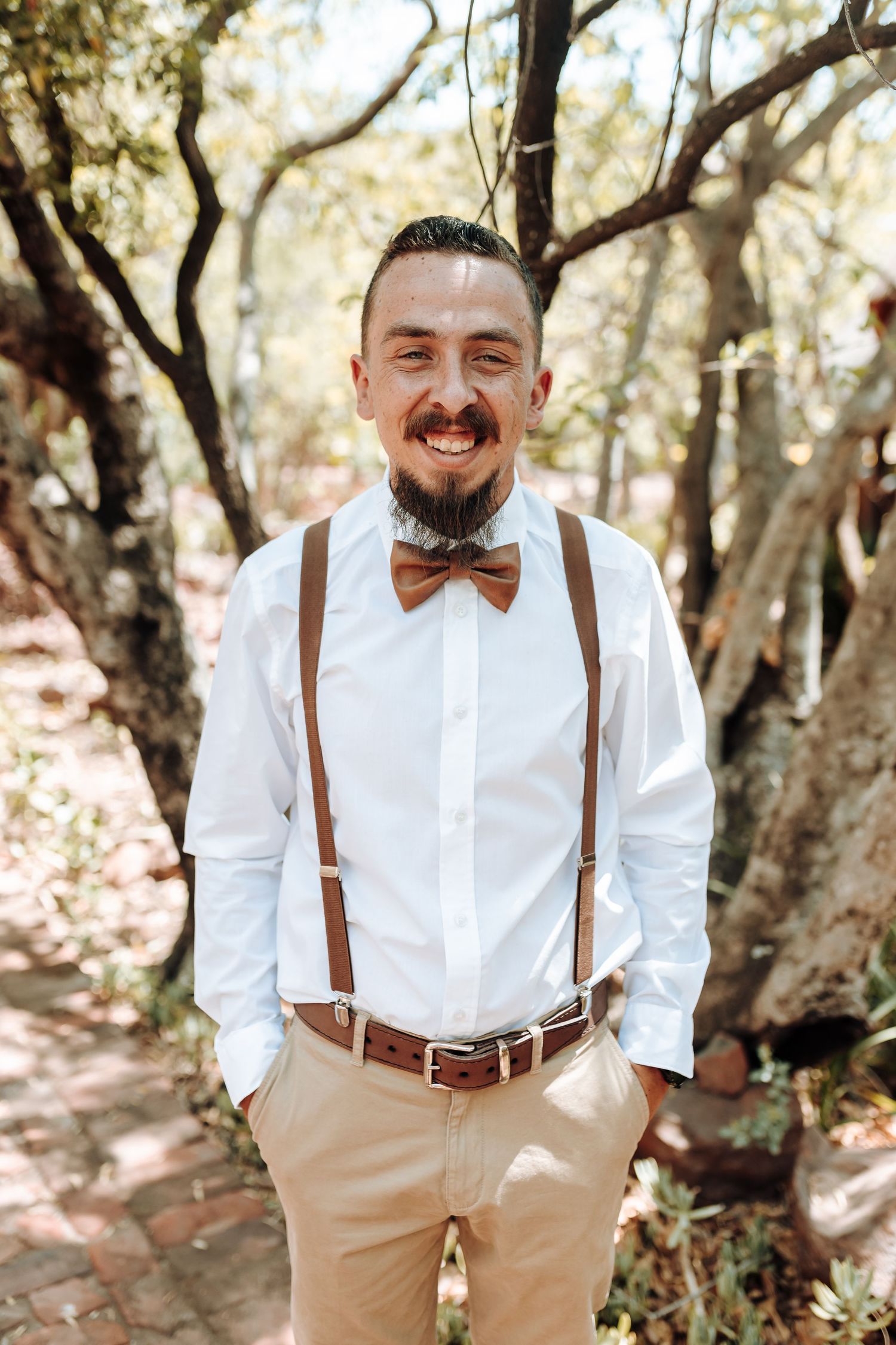 Groomsman wearing bow tie and brown suspenders poses confidently in woodland setting.