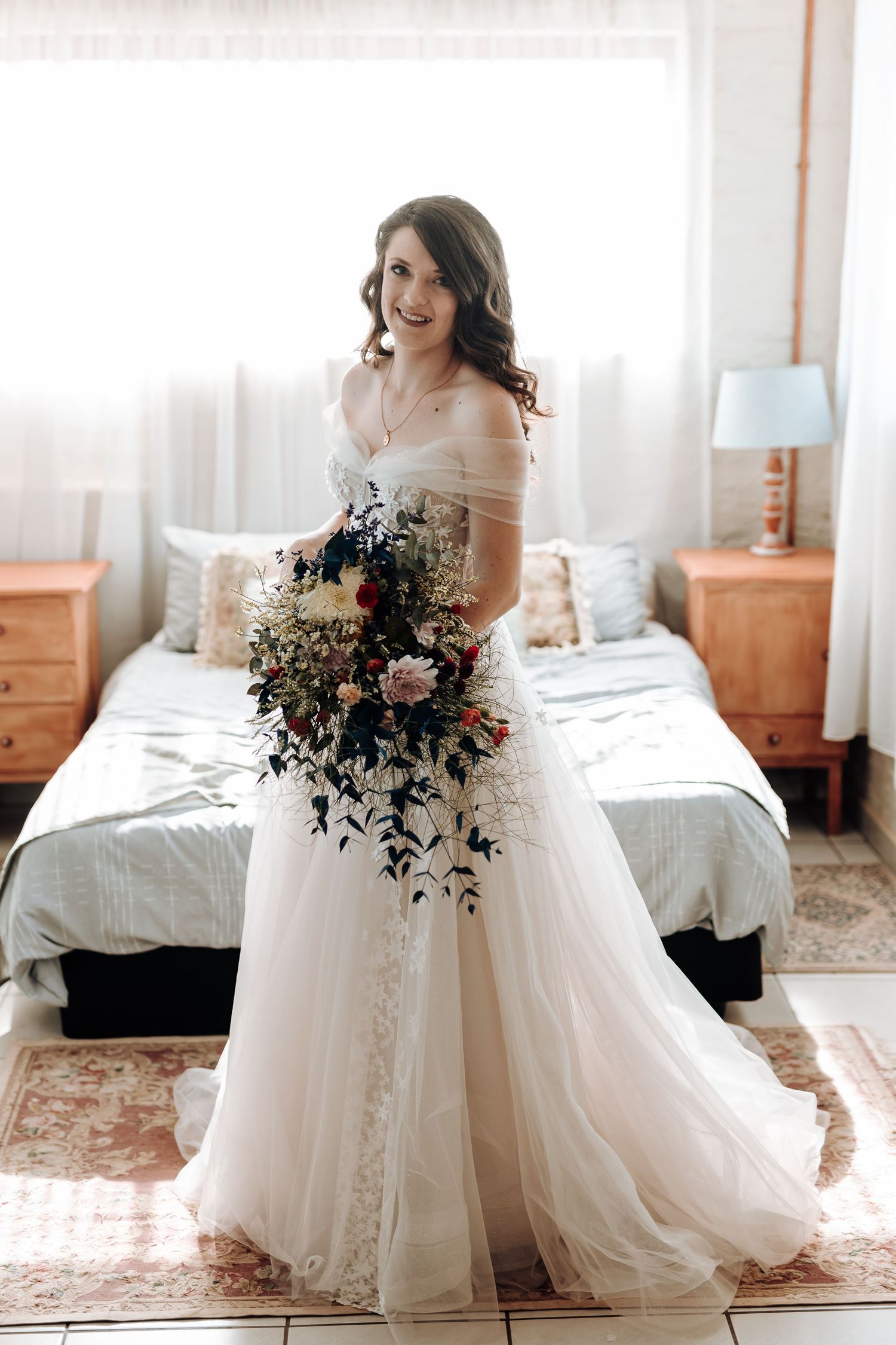 Bride in flowing tulle gown holding a cascading bouquet of mixed flowers in bedroom setting.