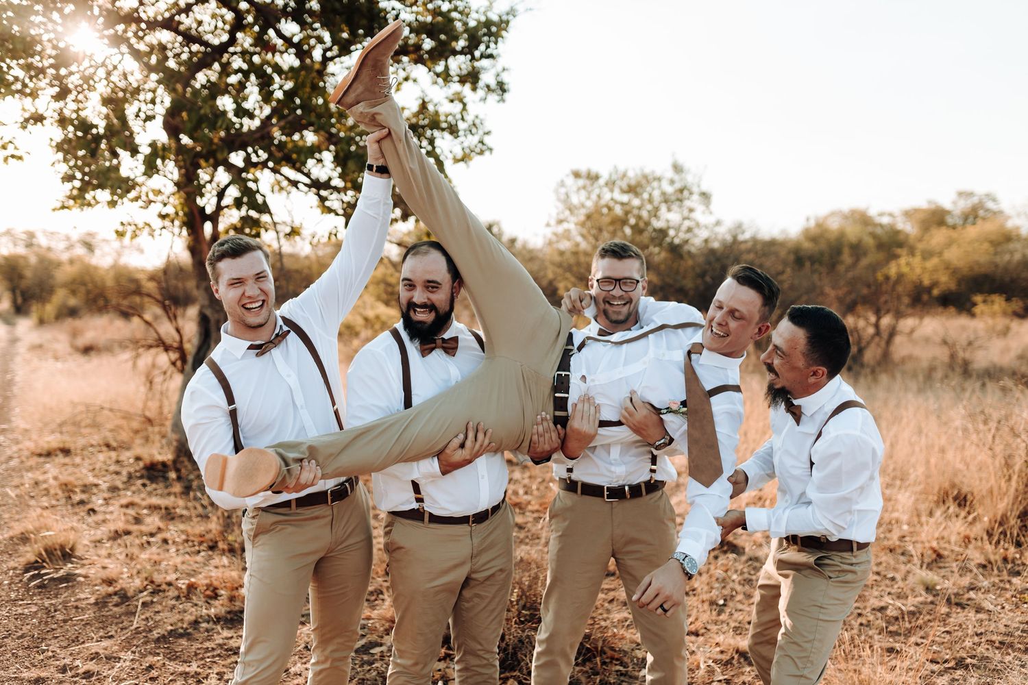 Wedding party in casual attire celebrates joyfully in a golden-lit rustic outdoor setting.