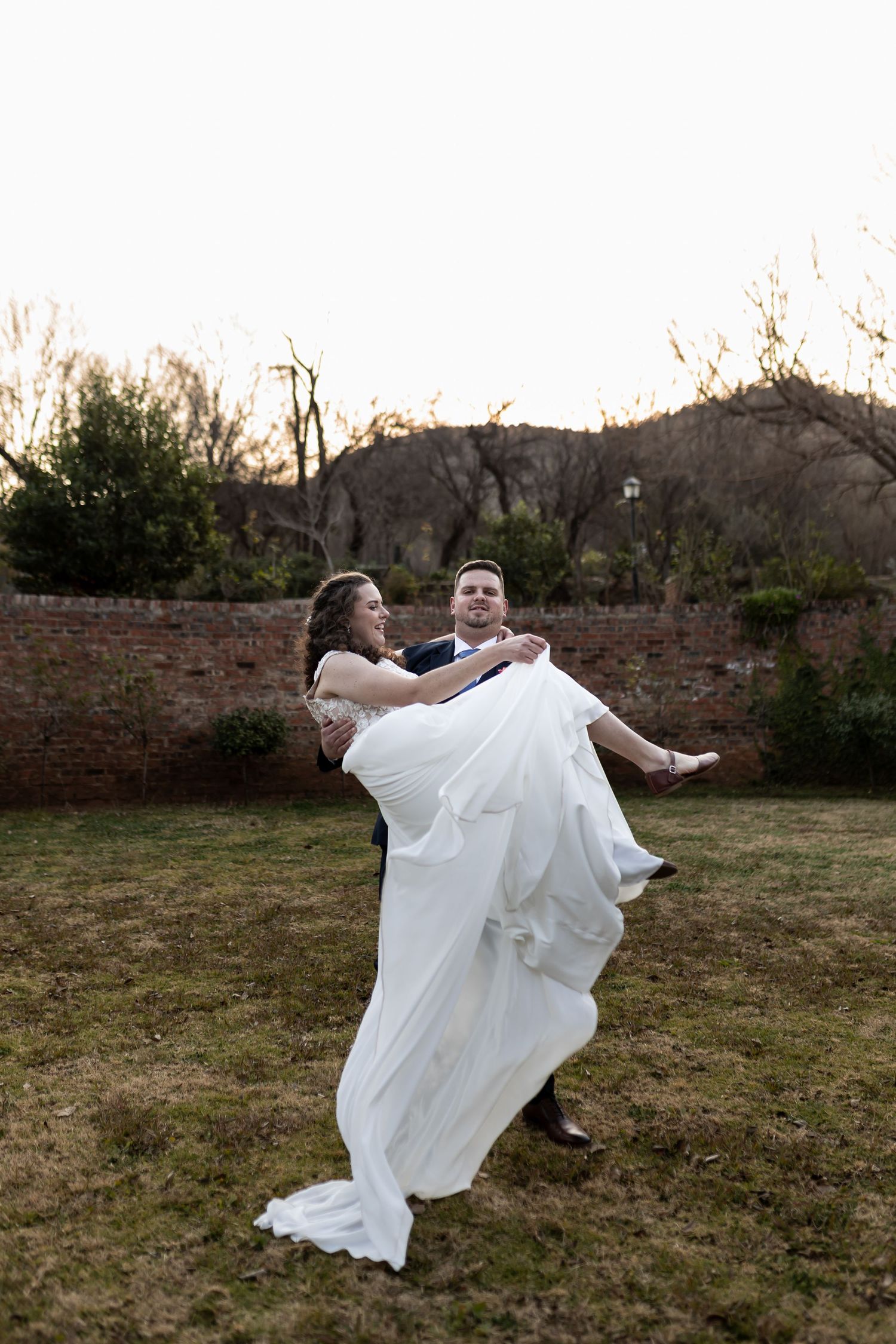 A couple in white wedding attire embraces while twirling on grassy lawn with brick wall in background at sunset.