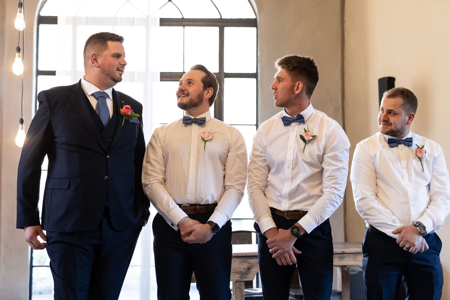 Groomsmen in formal attire stand together during a wedding ceremony at an elegant venue with arched windows.